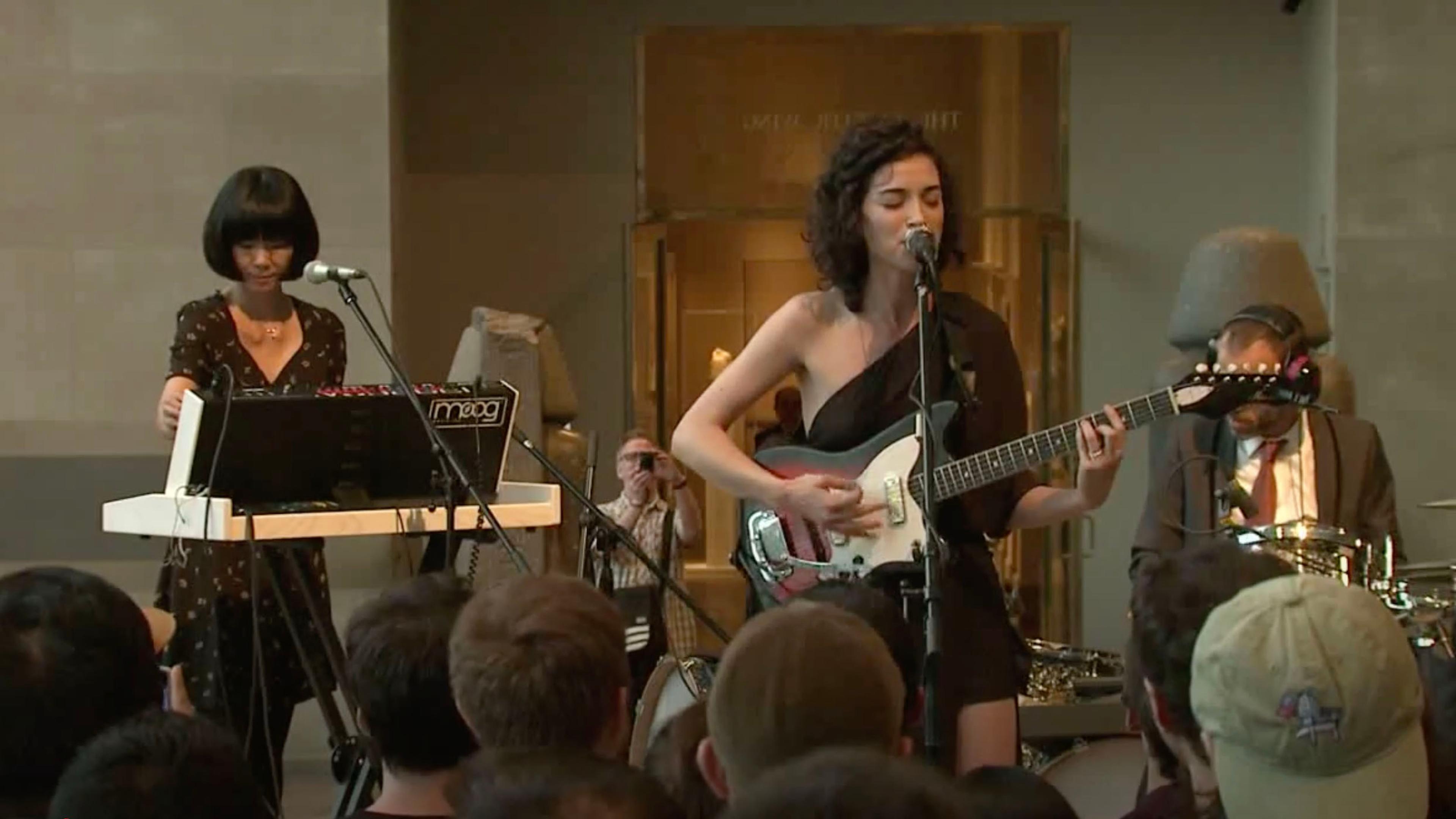 Three people performing music onstage; a woman plays keyboards on the left, a man plays drums on the right, and a woman stands center stage playing a guitar and singing