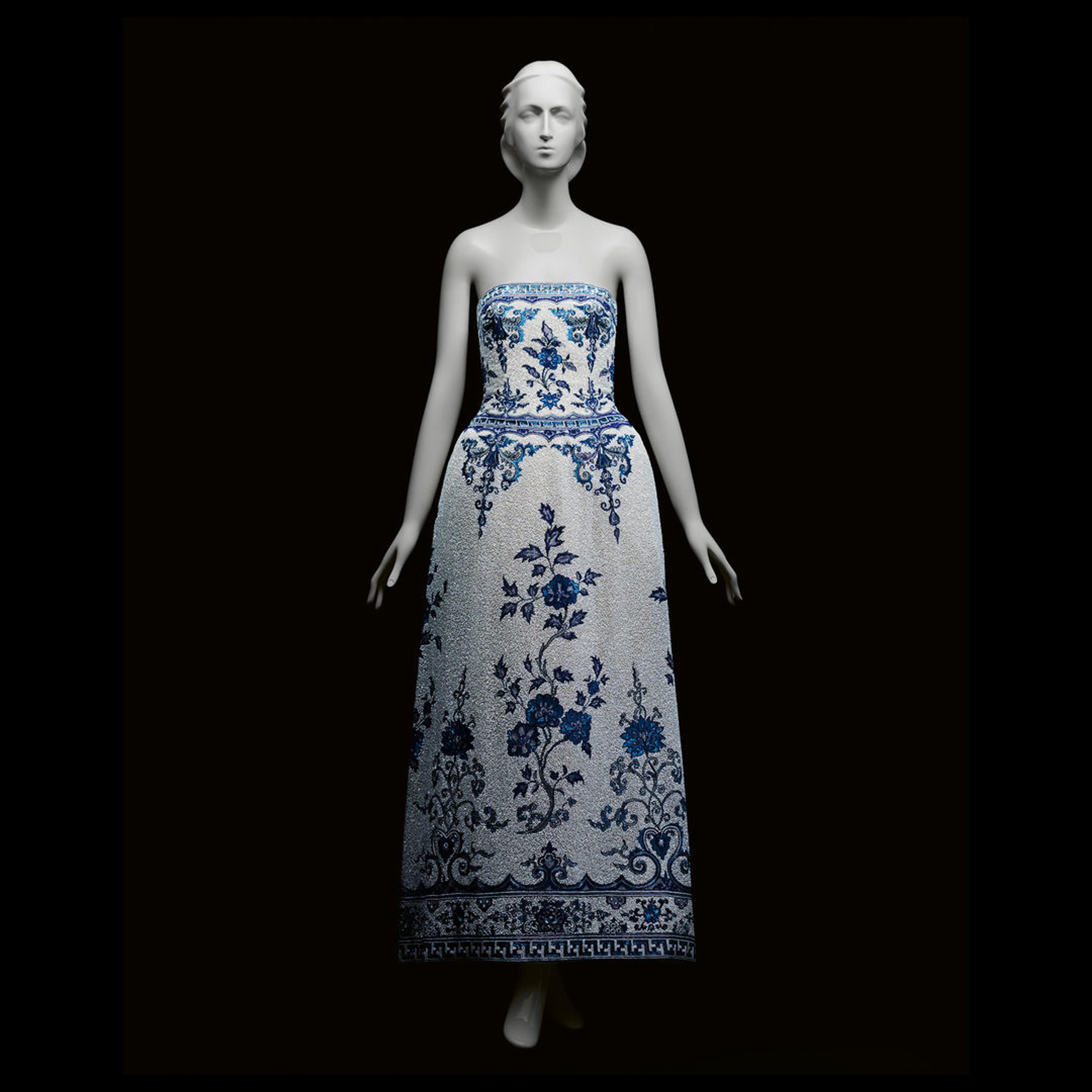 The back of a navy blue dress with beautiful white patterning on the back and lower part of the dress and around the wrists. The dress is against a grey background and fitted onto a mannequin with a head and hands.