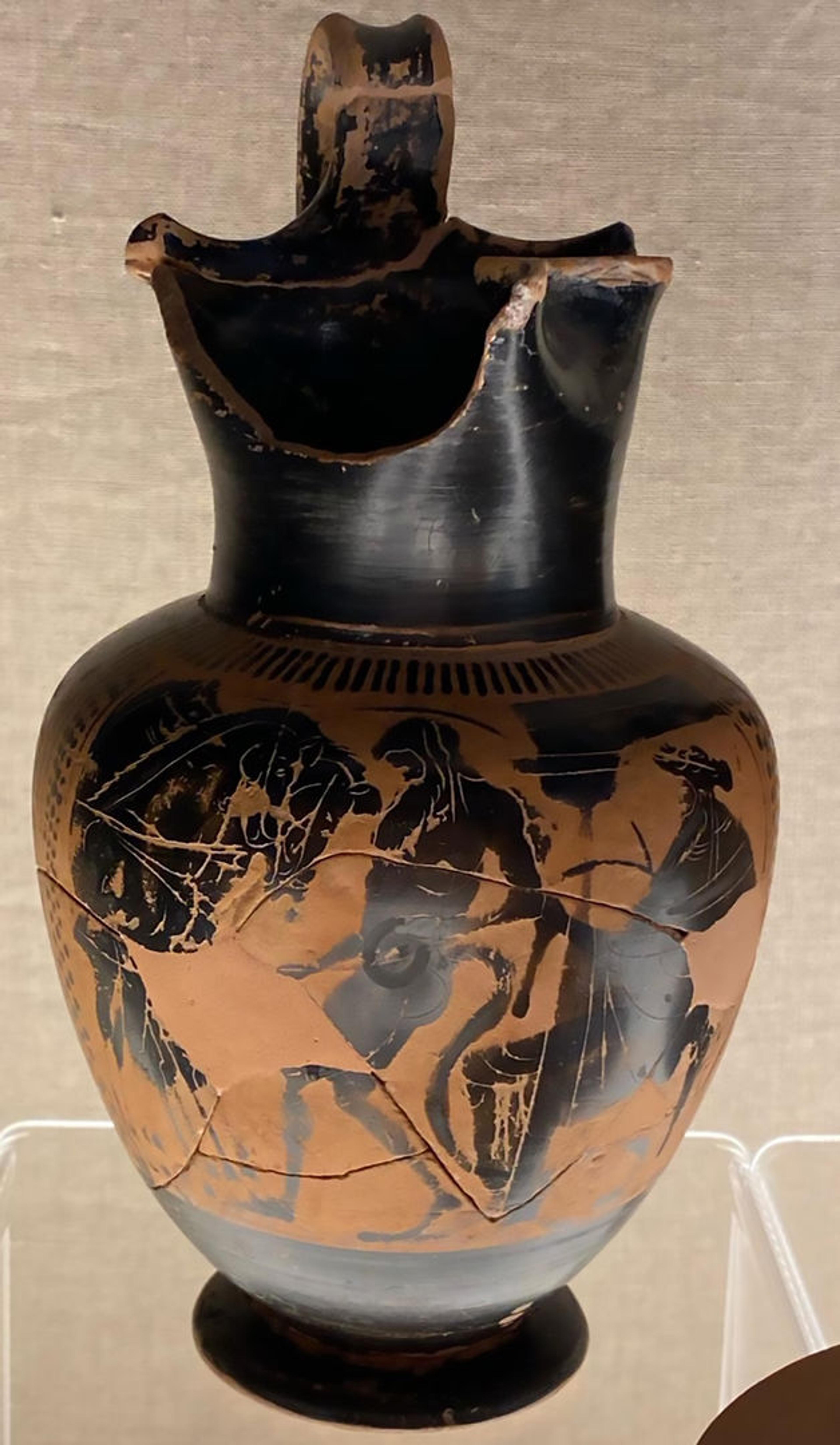 Black vase with a broken lip. The body of the vase depicts figures including Dionysos in black.