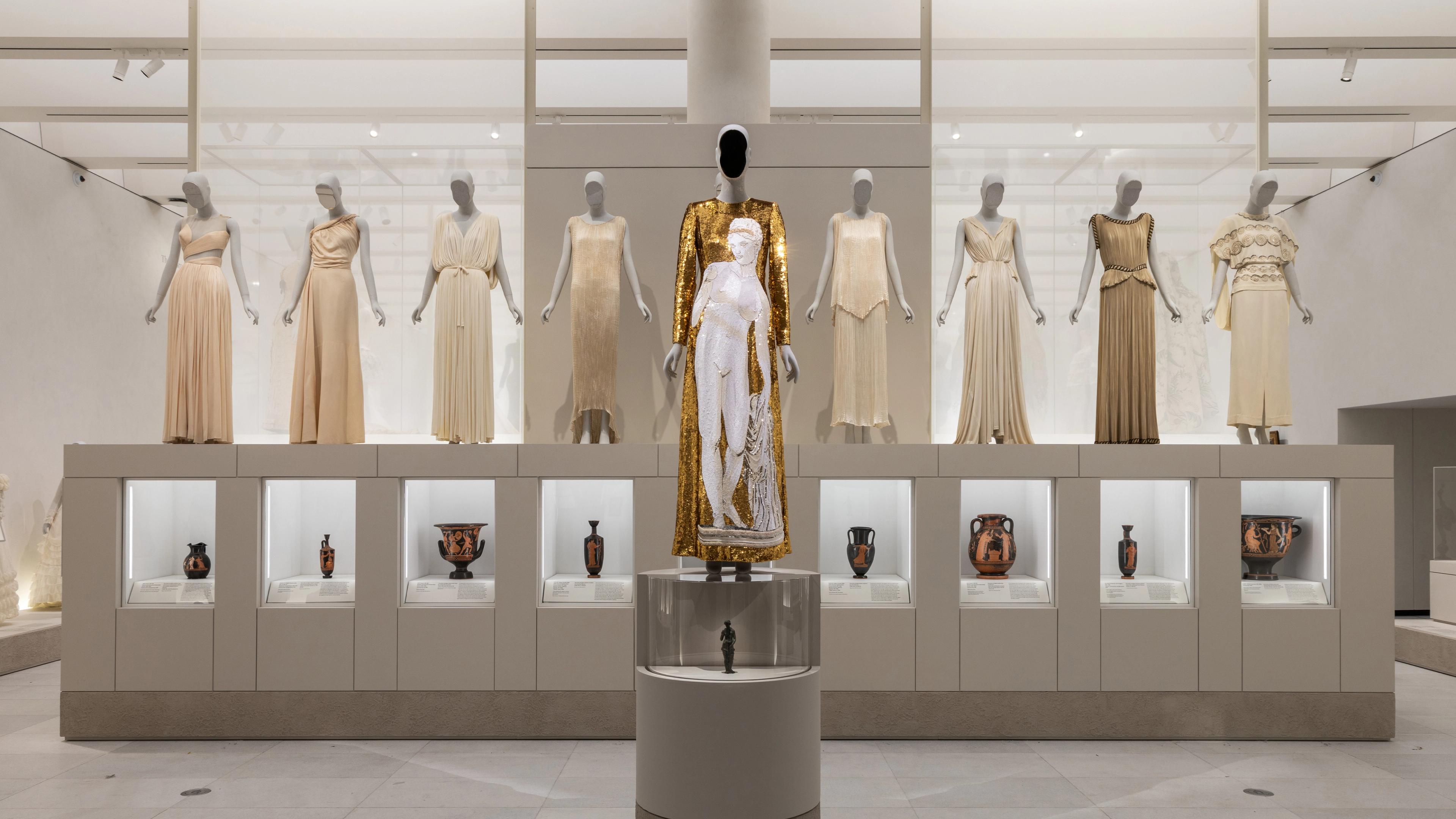 A museum exhibit displays ten elegant dresses on mannequins, with a central gold and white gown prominently featured. Below are decorative vases.