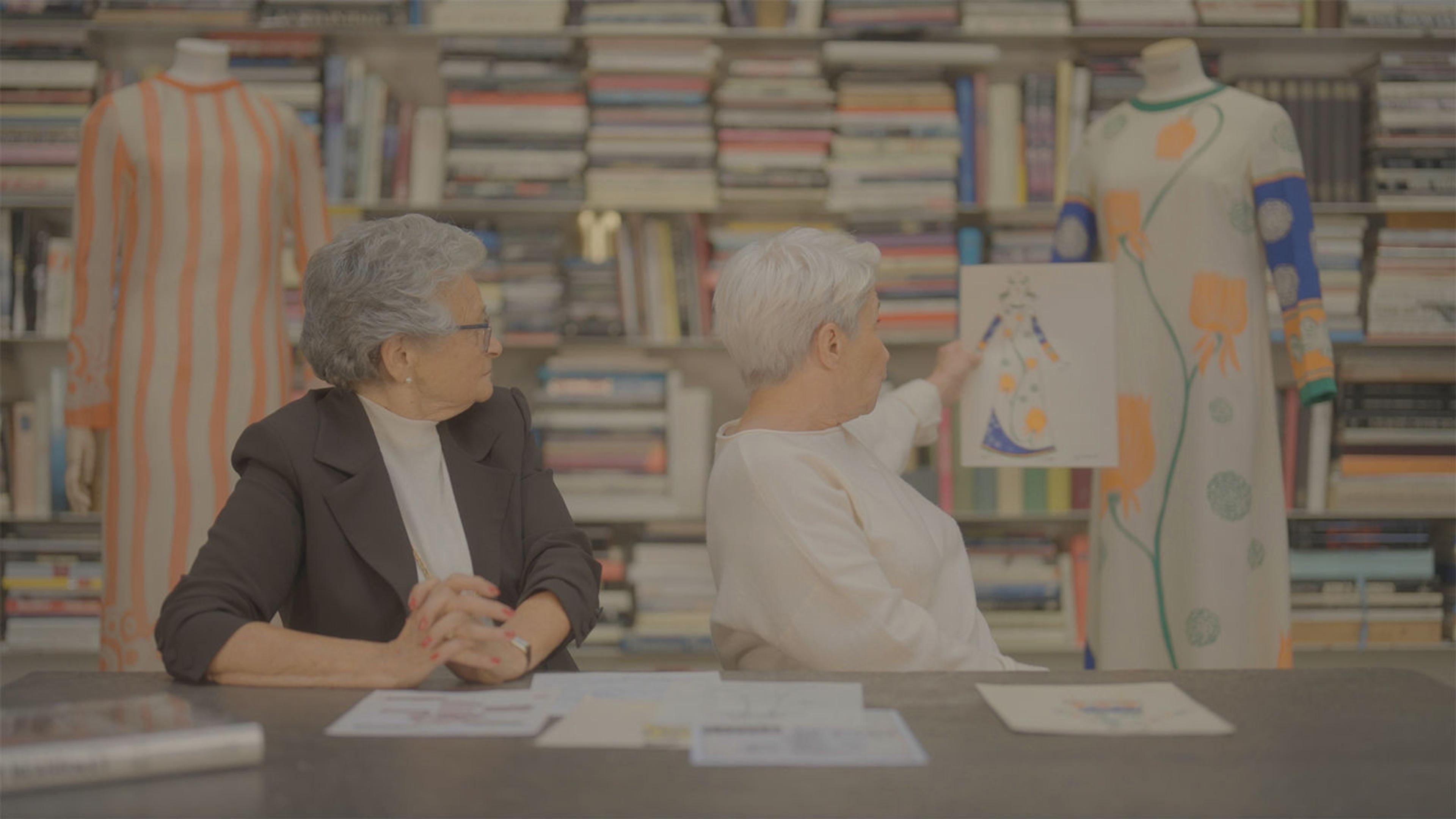 Two older women with short white hair in a studio whose background is a bunch of books. They are sitting at a wooden desk with papers in front of them. Both of their backs are turned looking at a dress in the background and the women to the right holds up a drawing of the dress.