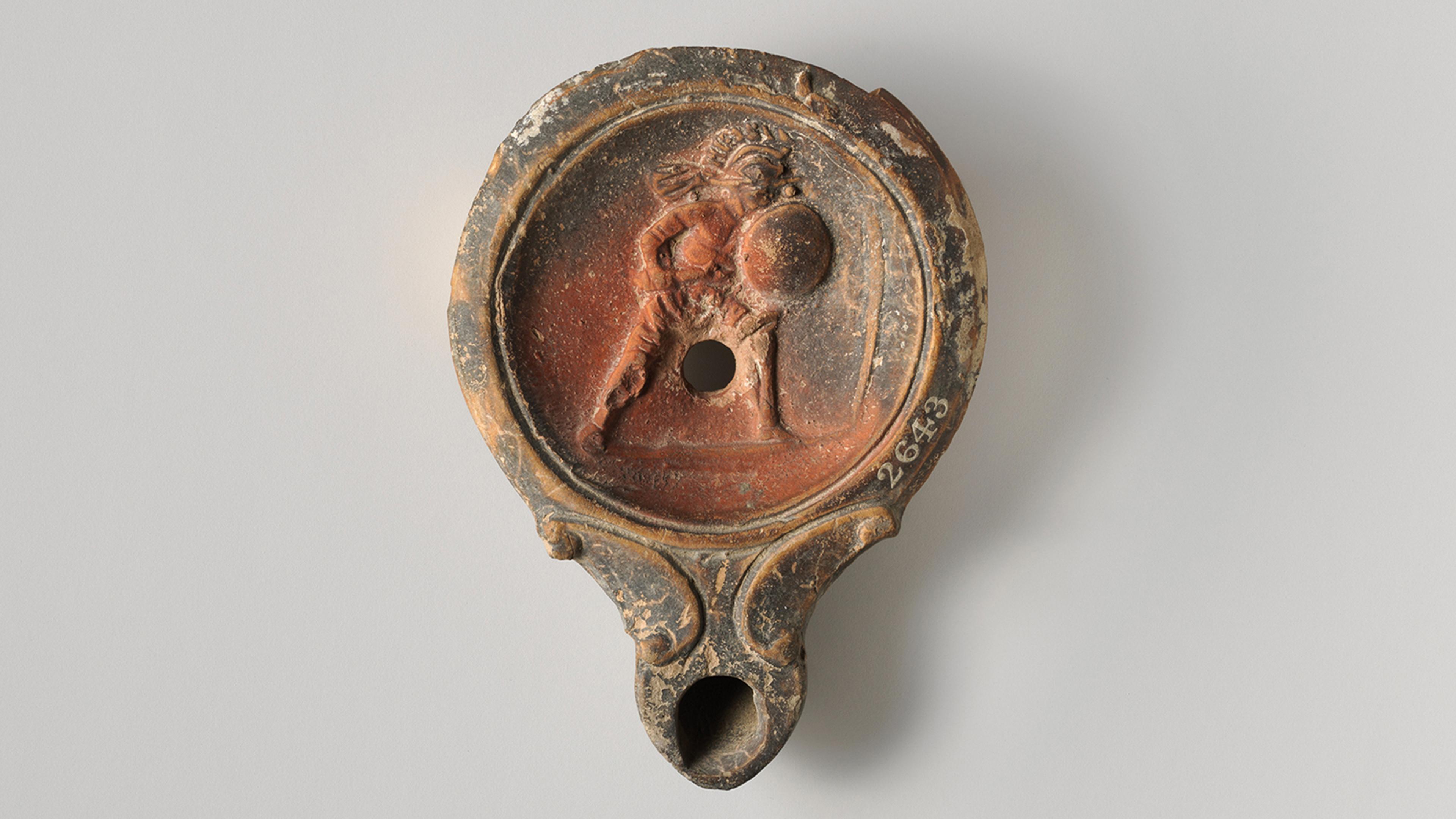Terracota oil lamp showing a gladiator, wearing a crested helmet, holding a round shield and short sword, advancing right.