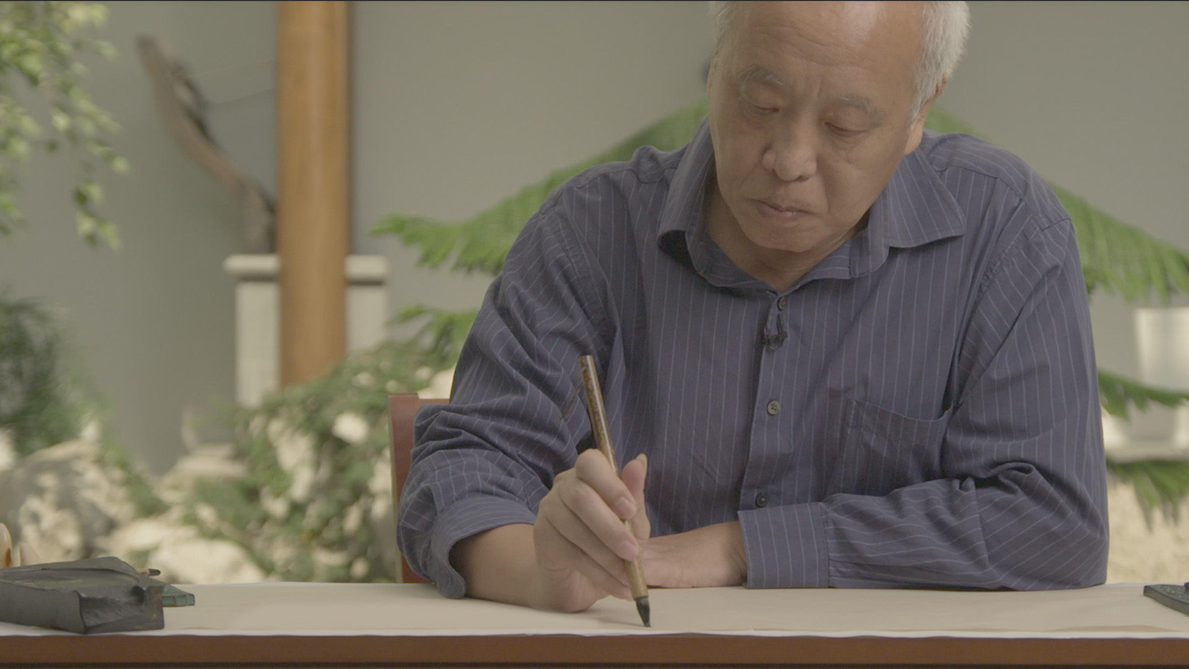 Image of an older Chinese man doing calligraphy with a brush and ink behind The Met's Astor Court with plants in the background.