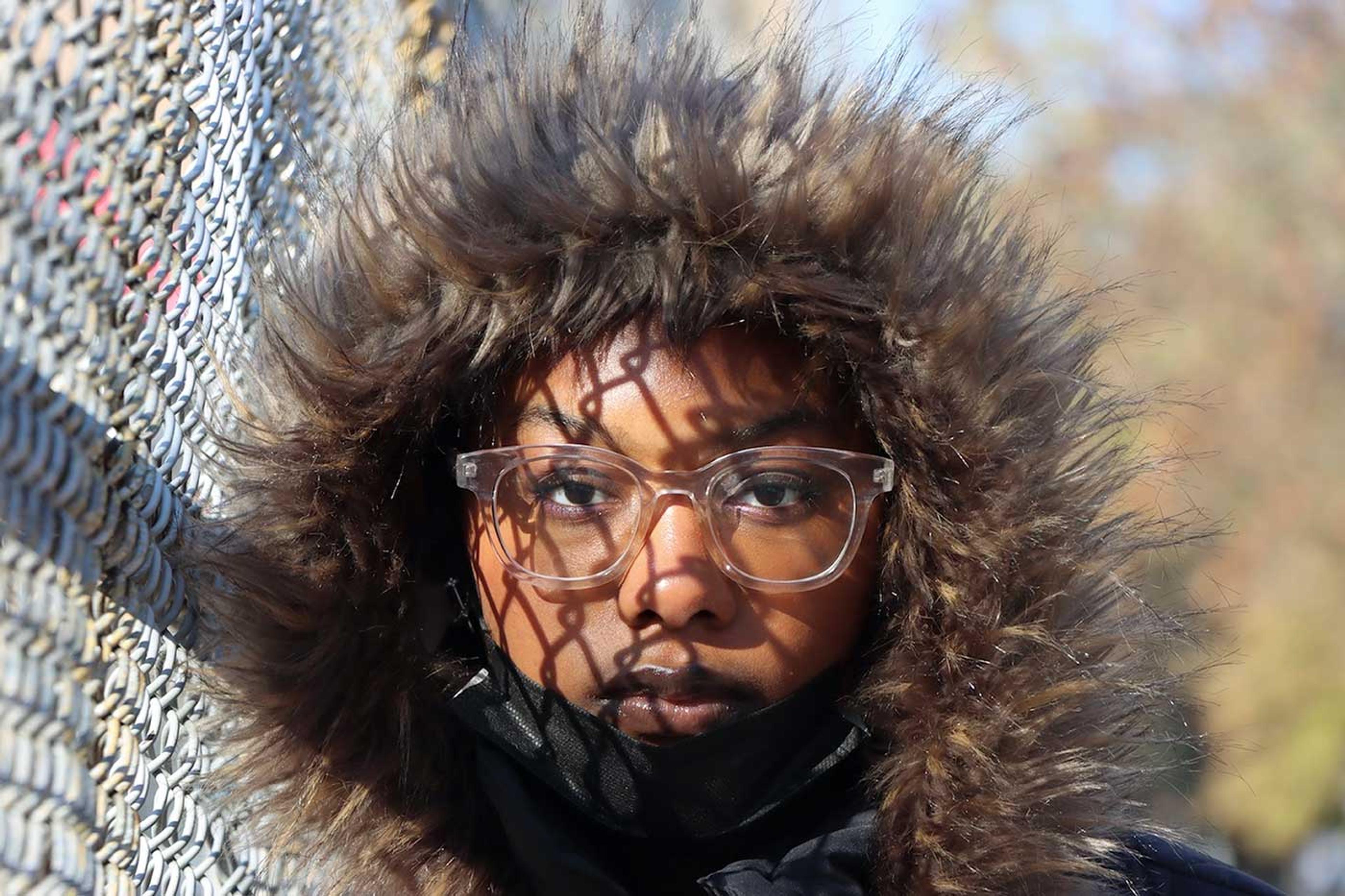 Photograph of a young girl in a hood standing next to a fence with shadows from the fence across her face.