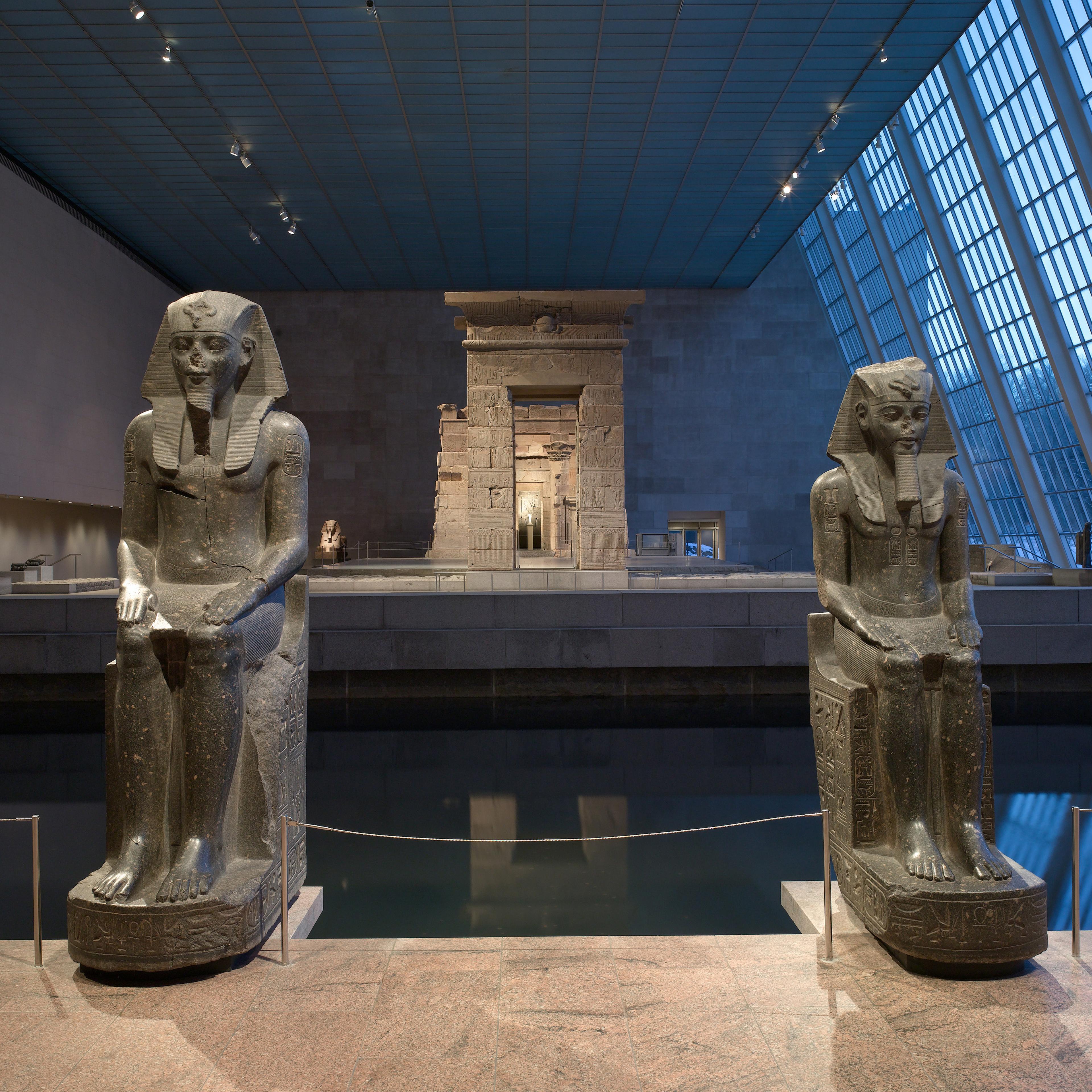 Two large seated statues of ancient Egyptian pharaohs flank a reflecting pool, with the Temple of Dendur in the background and light coming in from a tall wall of windows inside The Met.