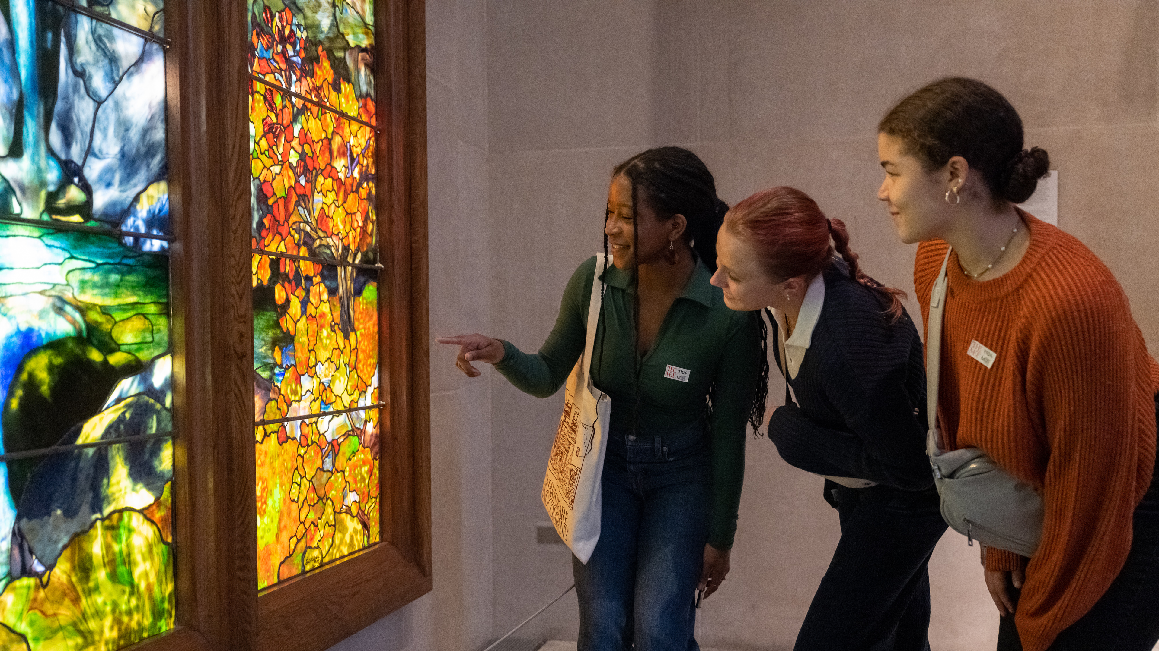 A group of teens look at an artwork in The American Wing at The Met. Design attributed to Agnes F. Northrop (1857–1953); made by Tiffany Studios (1902–32). Autumn Landscape, 1923–24. Made in New York. Leaded Favrile glass, 132 x 102 in. (335.3 x 259.1 cm). The Metropolitan Museum of Art, New York, Gift of Robert W. de Forest, 1925 (25.173a–o)