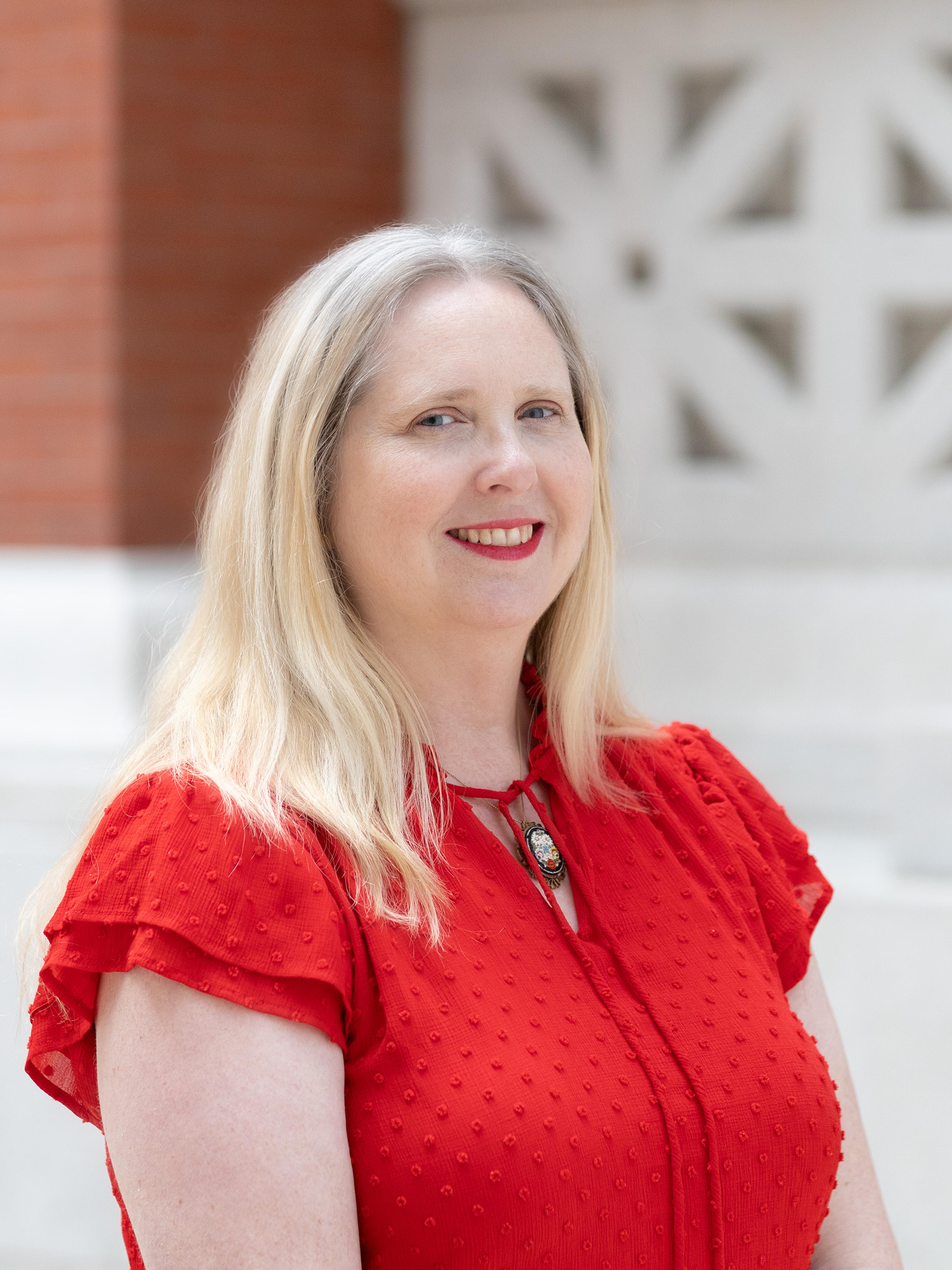 A woman wearing a bright red blouse smiles in front of a brick background