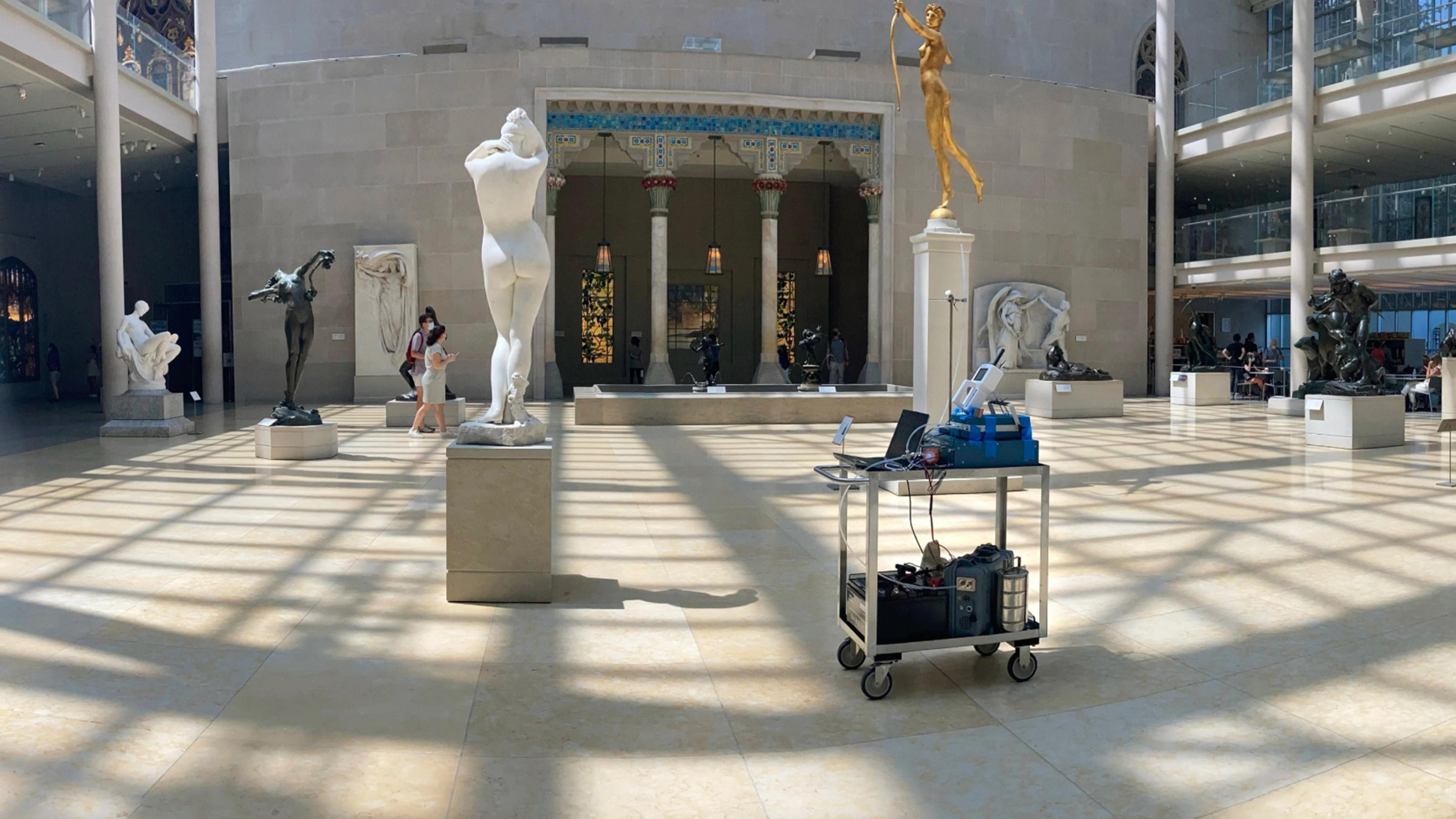 A cart on wheels with scientific machinery is parked in the middle of the sunlit American Wing sculpture courtyard; a grid of shadows from the overhead skylights creates a grid pattern on the polished stone floors.
