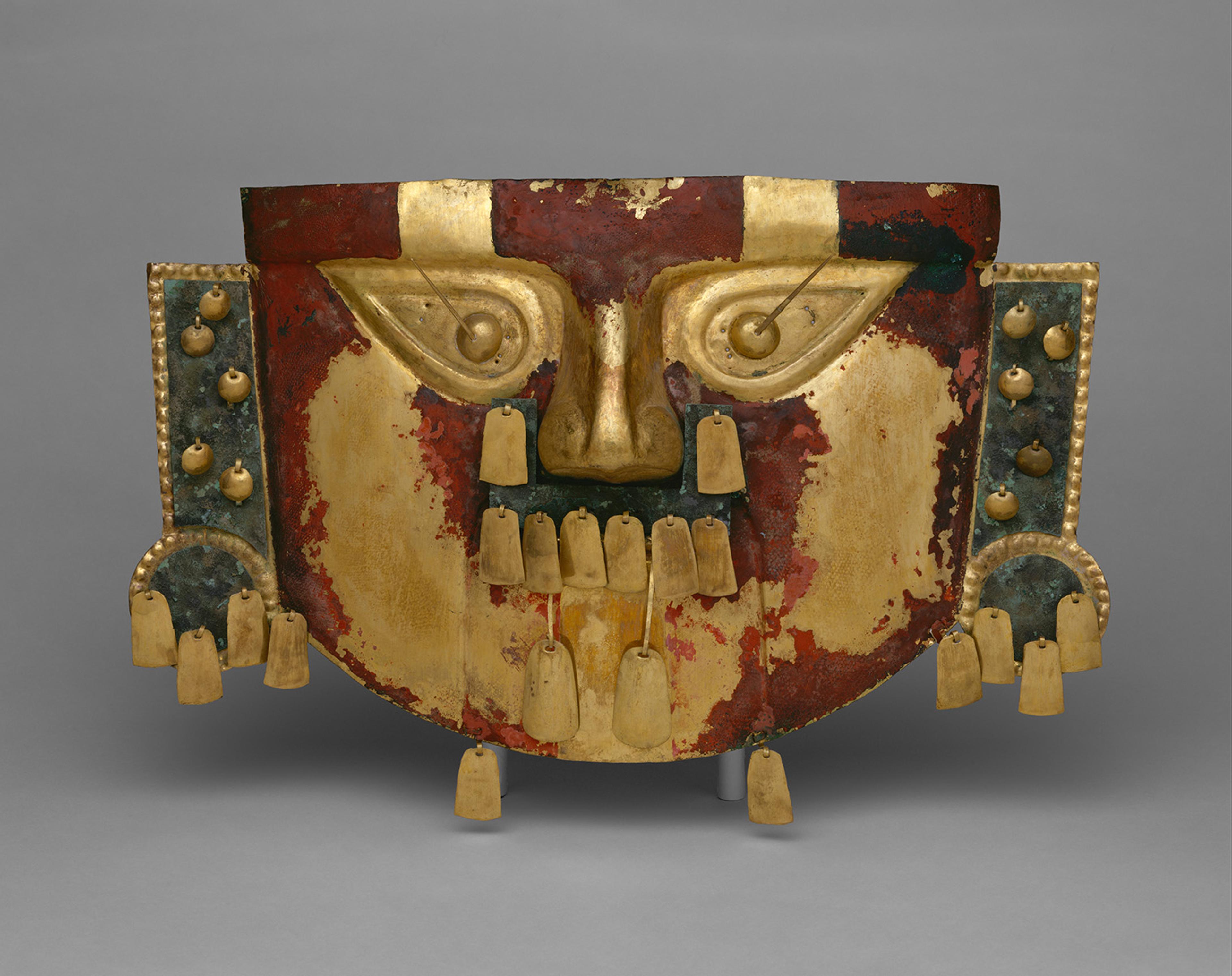 Ancient ceremonial mask with a weathered red, gold, and black face, wide eyes, dangling ornaments, and side panels