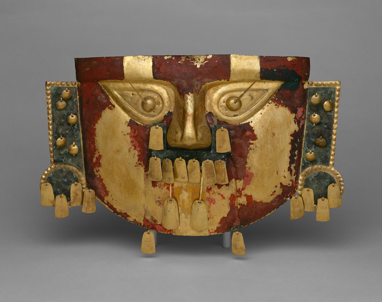 Ancient ceremonial mask with a weathered red, gold, and black face, wide eyes, dangling ornaments, and side panels