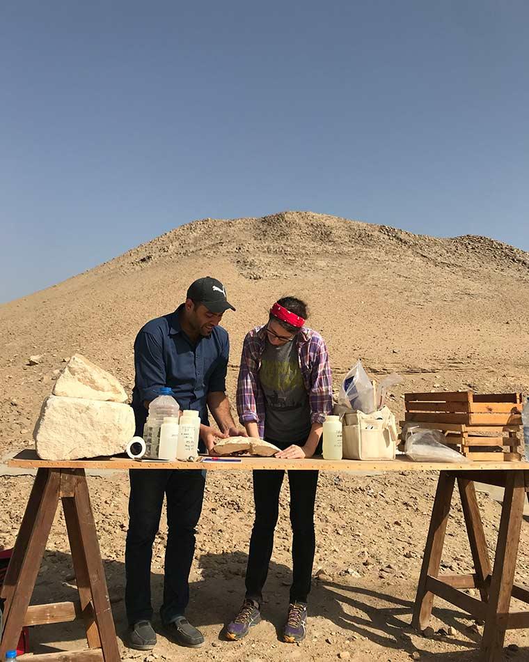 Two people examining an object while standing at a makeshift table in the desert.