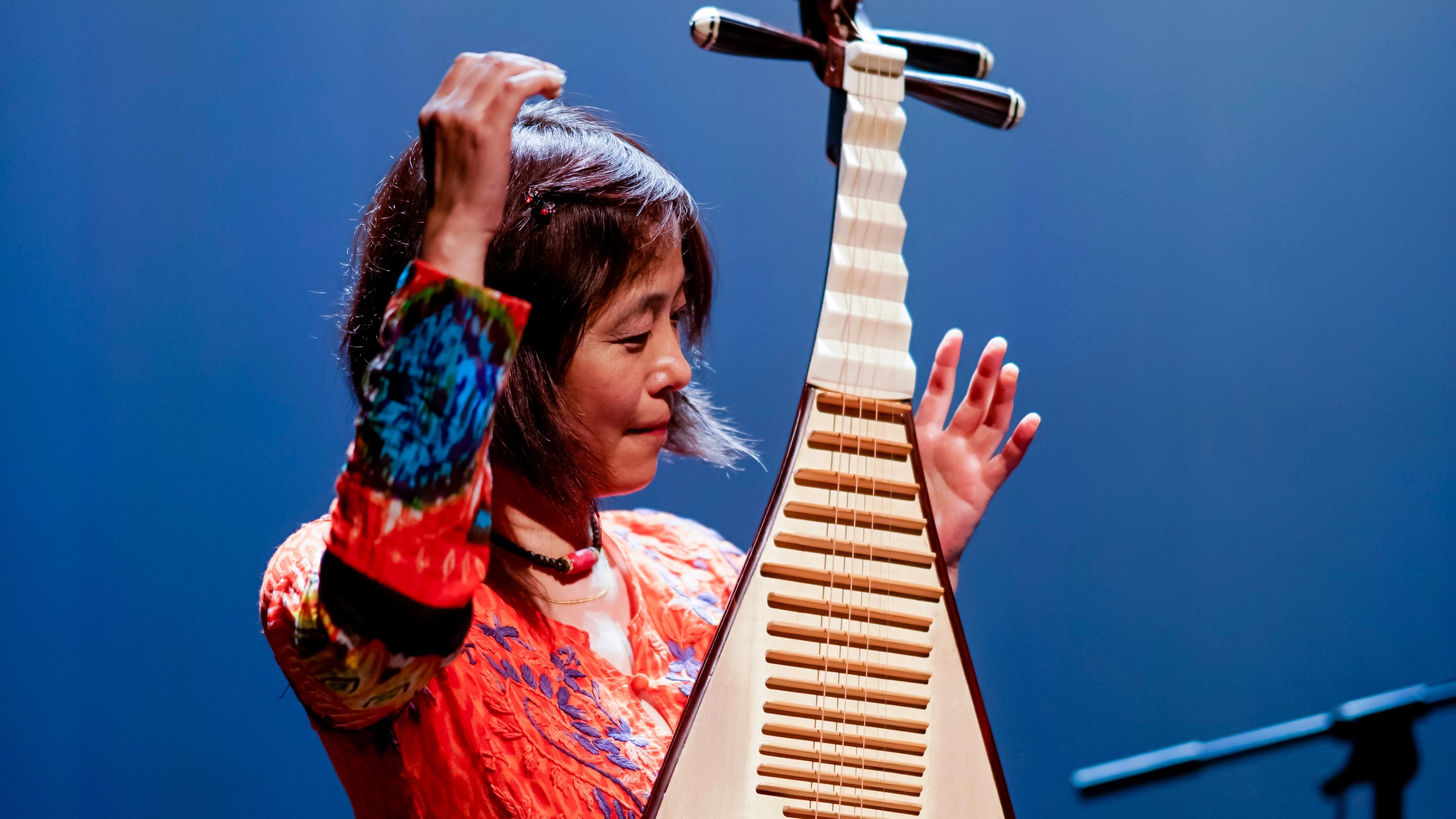 Wu Man, hands aloft, playing the pipa on stage against a deep blue background.