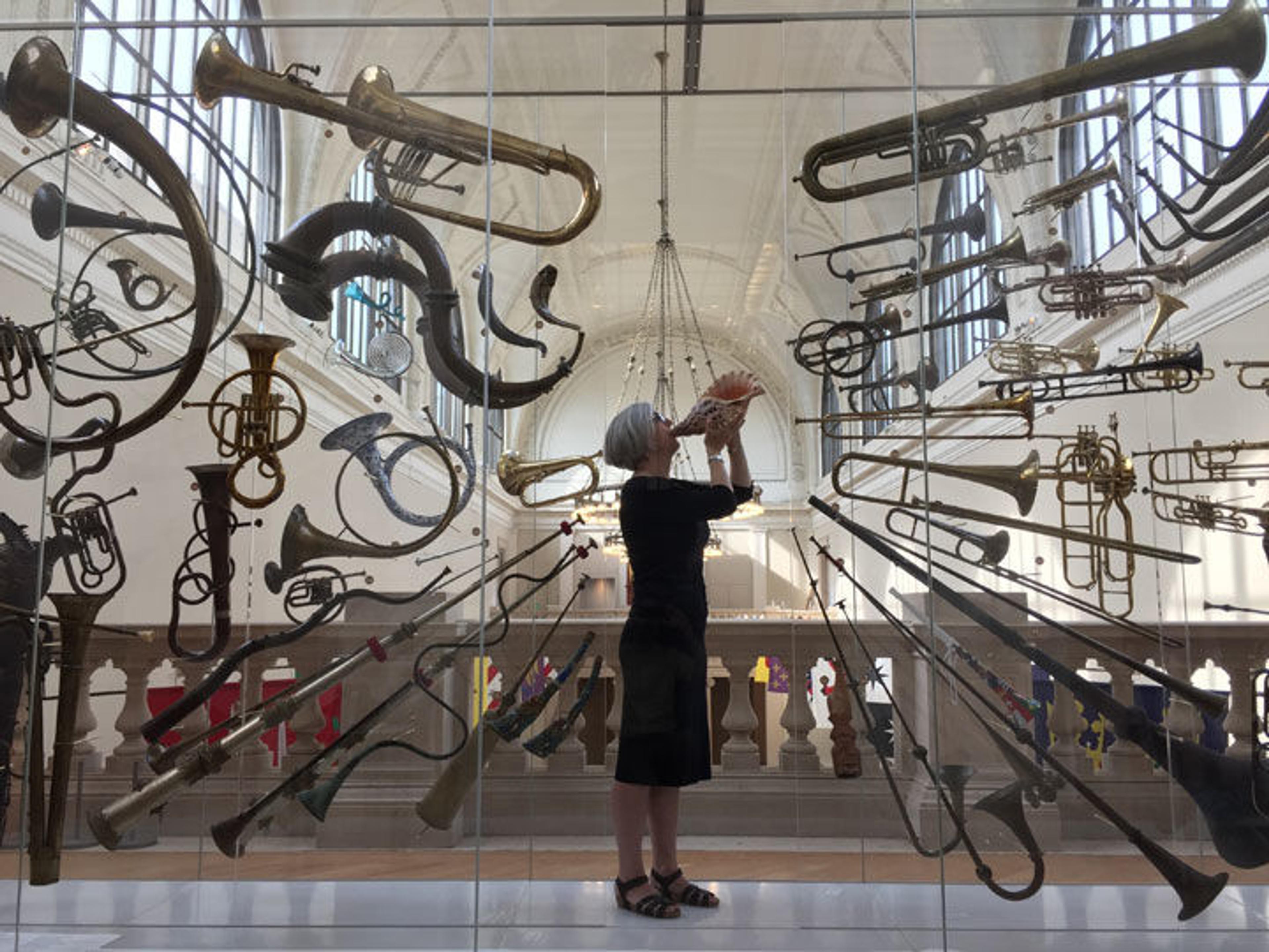 The author stands among a large display of brass instruments while blowing into a conch shell