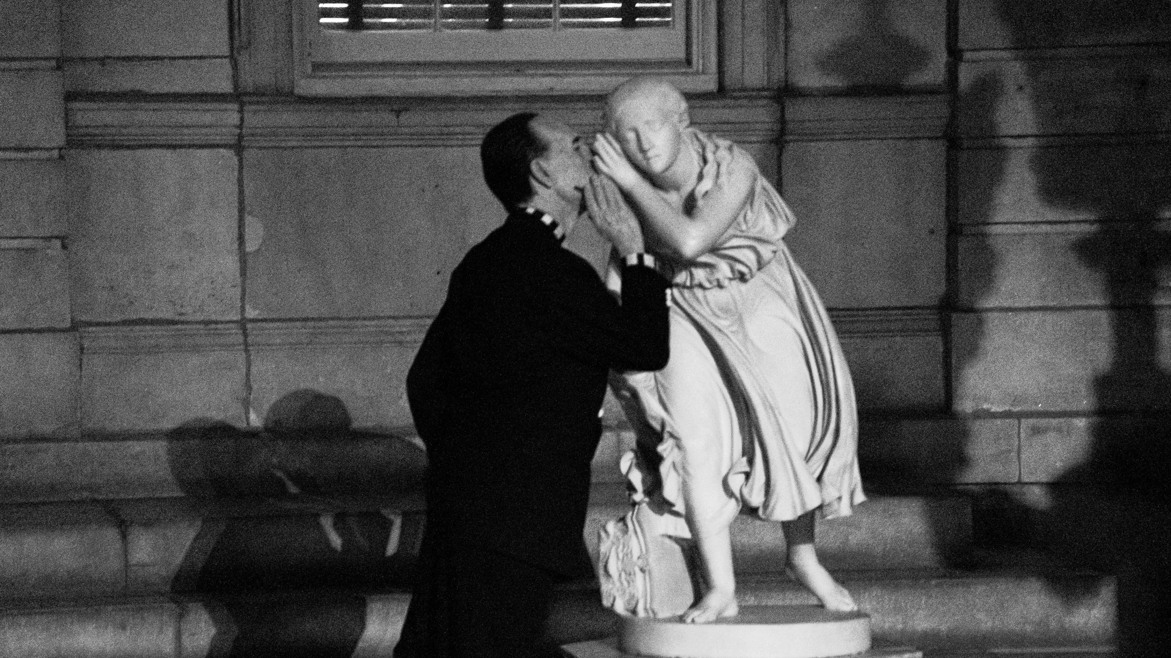 Black and white photograph of a man in a long black coat whispering in the ear of a marble sculpture of a young woman who lifts her hand to her ear as if to hear what he is saying.