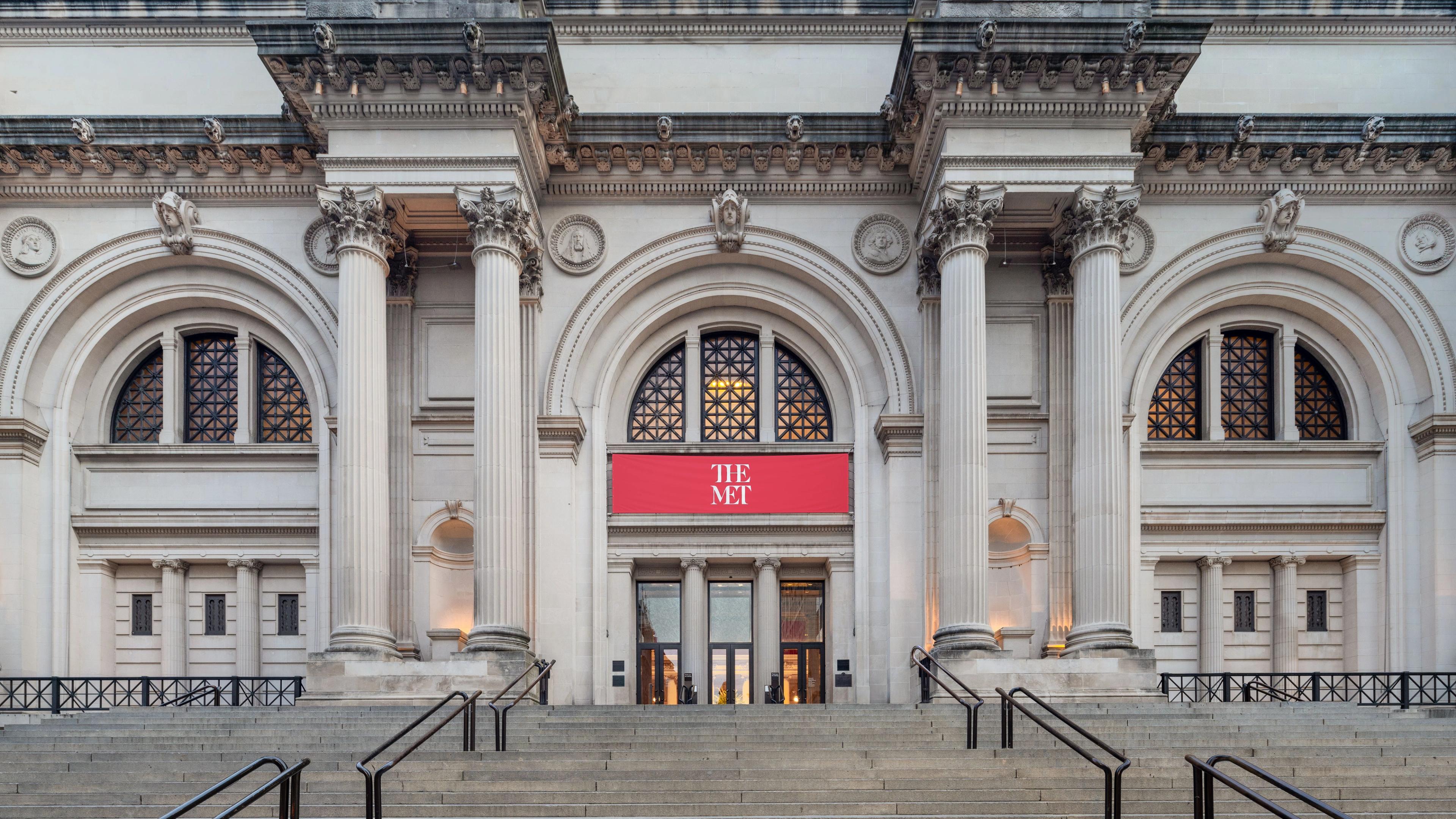 The Metropolitan Museum of Art's fifth avenue entrance on a clear day featuring tall columns and a prominent red banner displaying "THE MET."