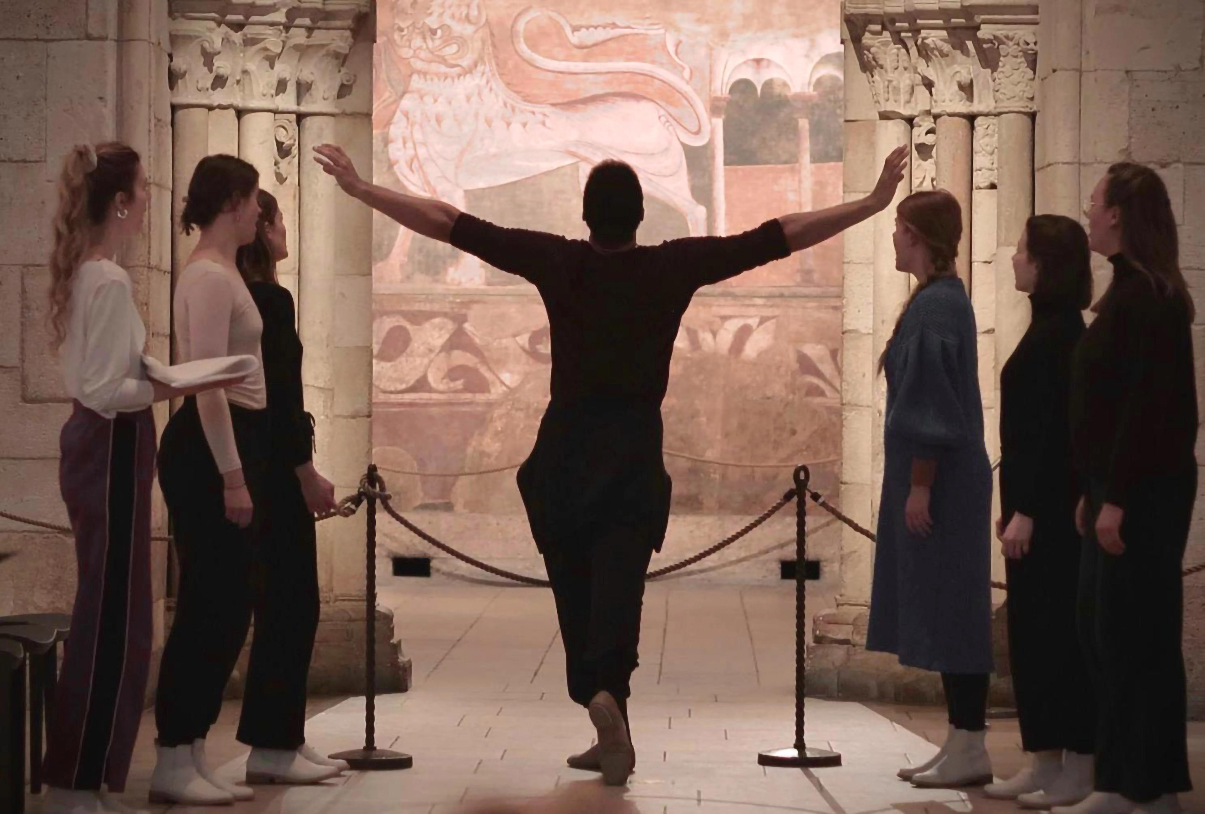 Dancer with their arms outstretched, facing a doorway through which a fresco is framed. Three figures stand on either side of the dancer.