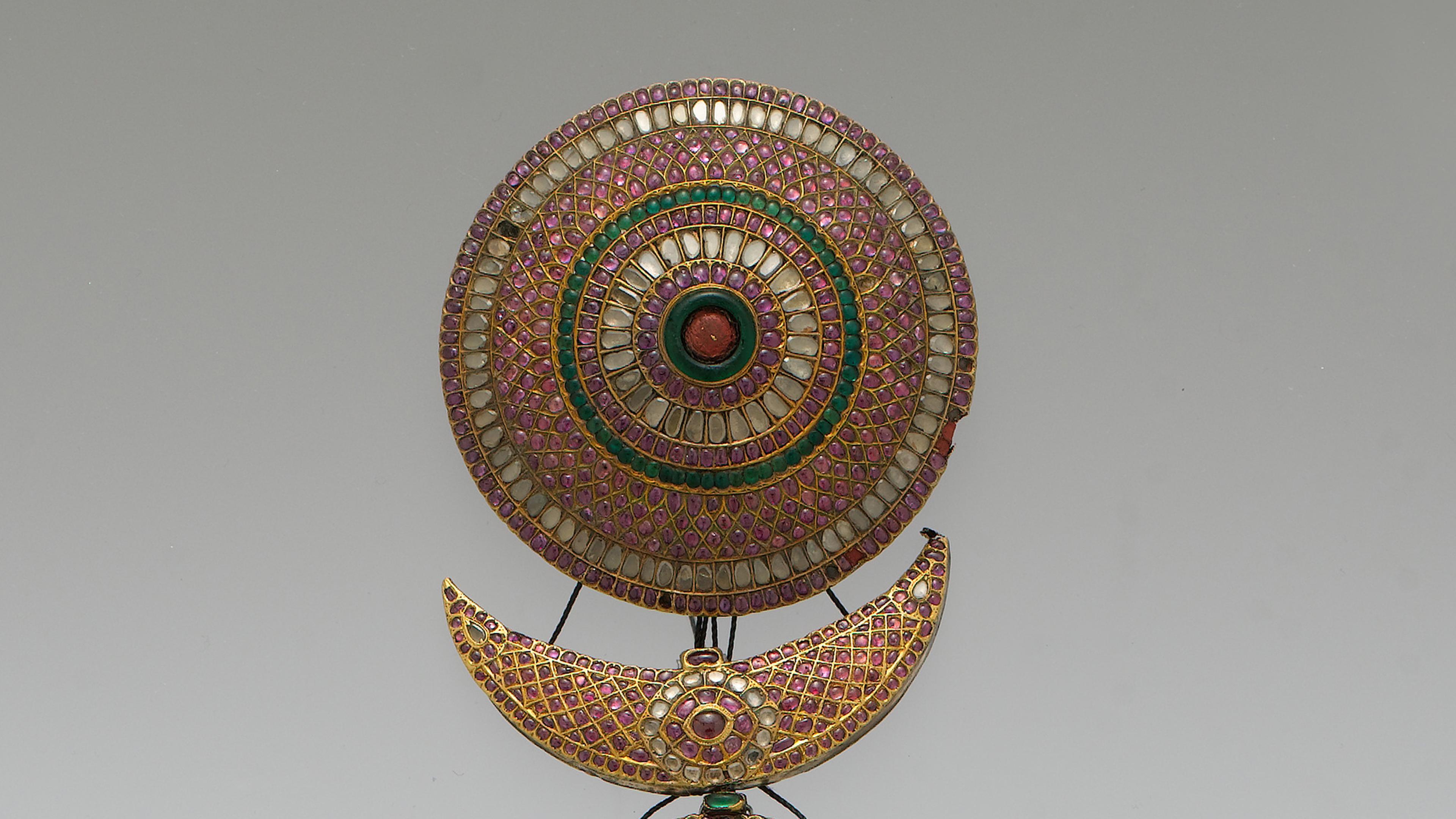 Close up of a plait ornament accented with brightly colored stones. The top part of the ornament is round, and hanging underneath is a crescent-shaped piece.