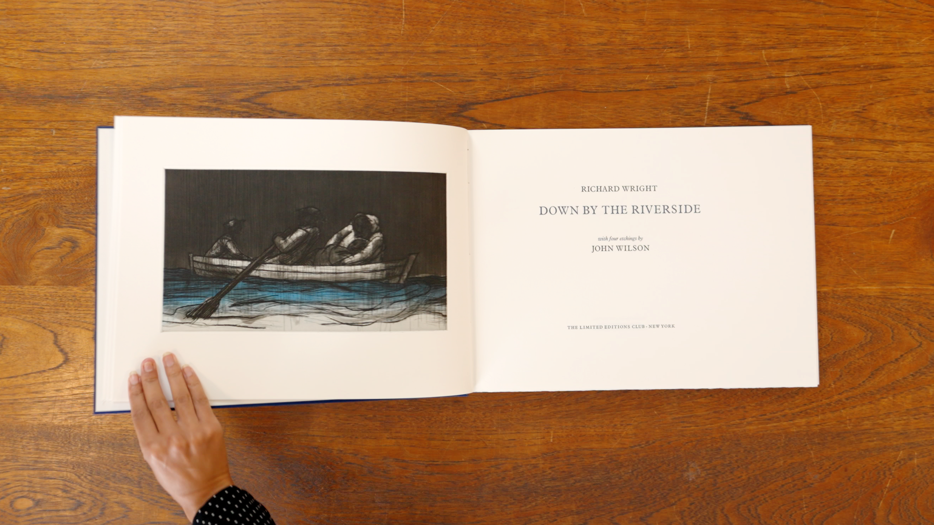 A book presented on a wooden table, with a hand on the left side of the page preparing to turn the page.
