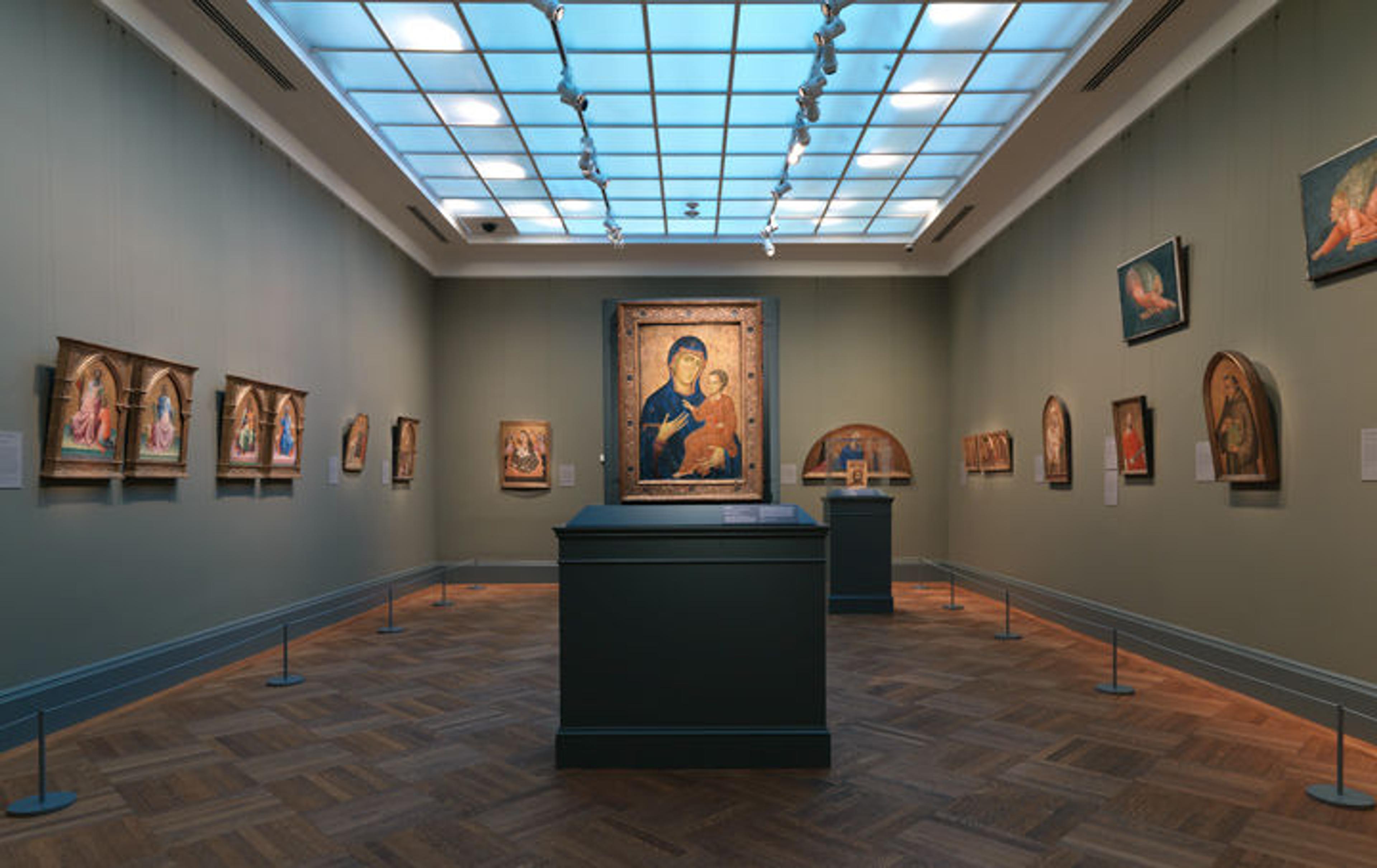 A serene gallery with European Paintings hung on the walls in gold frames and a large painting in a pedestal display in the middle of the room. Natural sunlight from a glass ceiling illuminates the room. There are no people in the gallery.