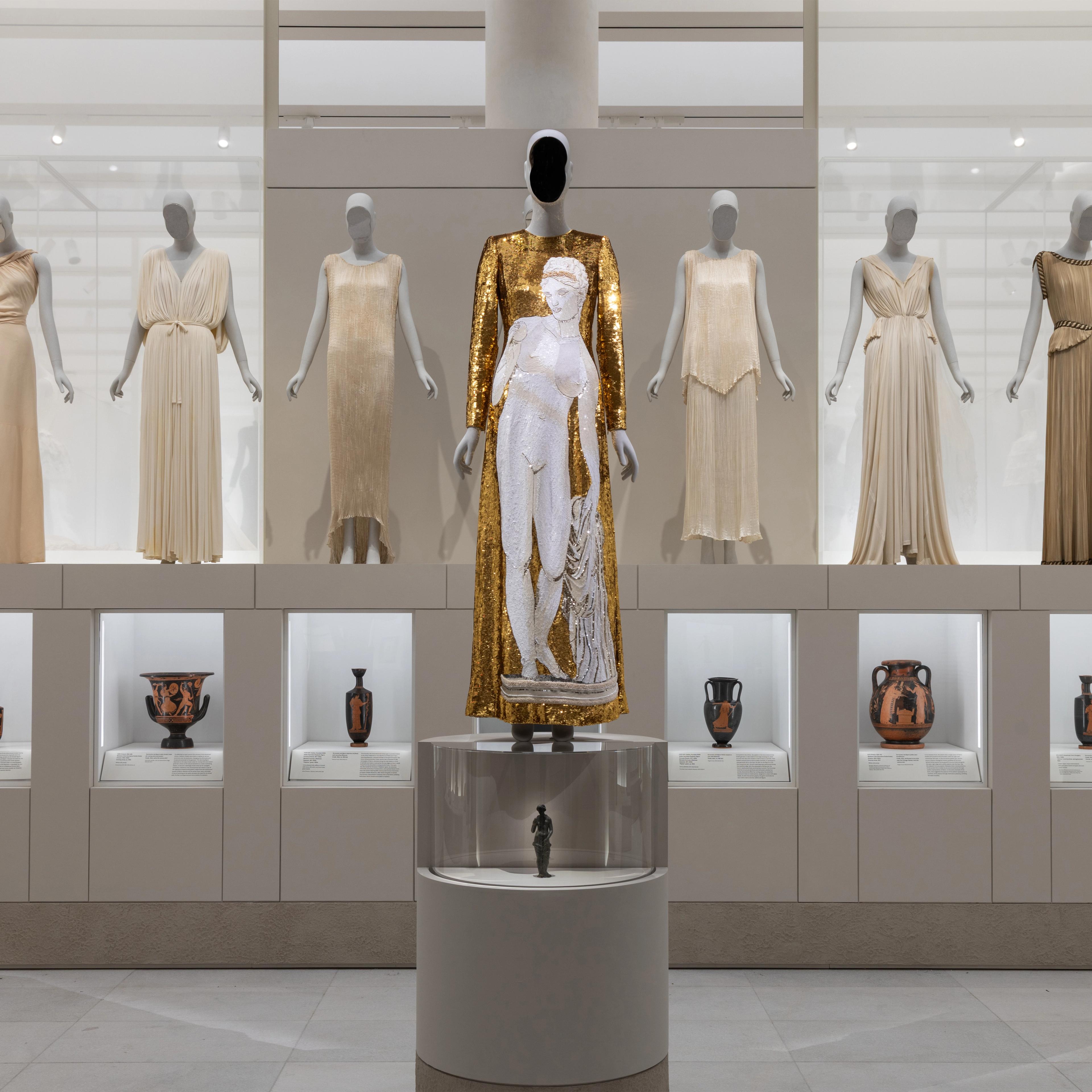 A gallery display features mannequins dressed in elegant gowns, with a central mannequin in a gold and white outfit, surrounded by ancient pottery in glass cases below.
