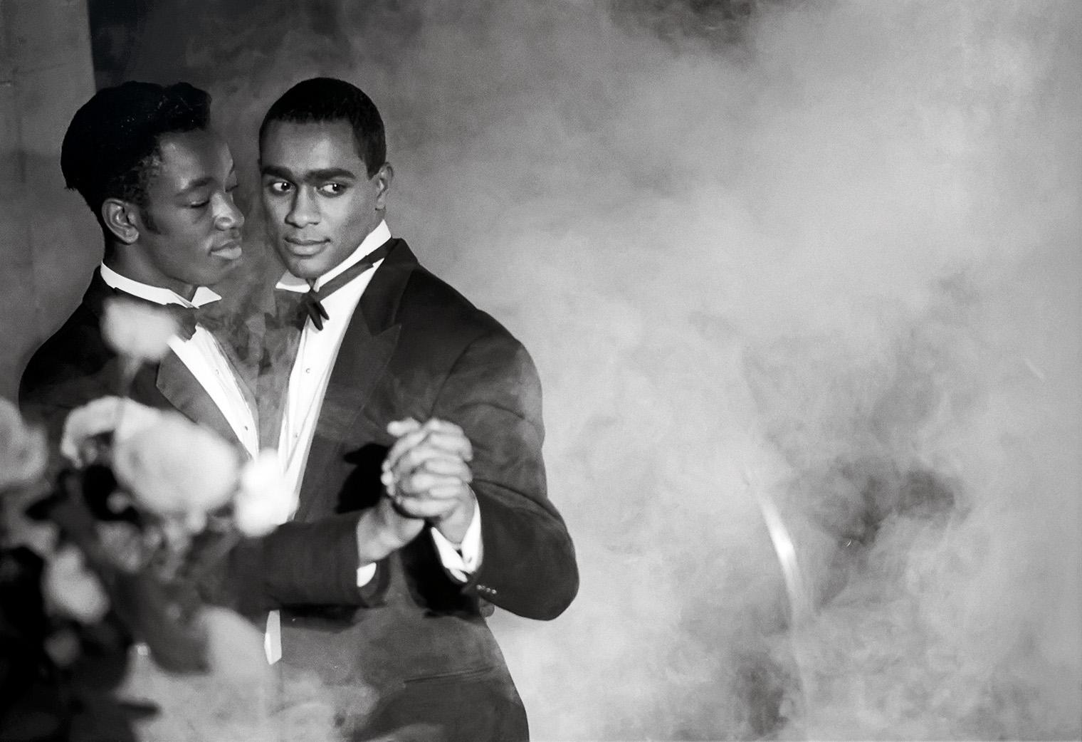 Two men in tuxedos dance closely, enveloped in soft, swirling fog. The scene conveys intimacy and warmth, with a romantic and serene atmosphere.