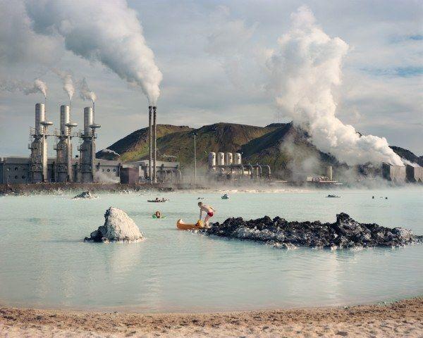 Color photograph by Laura McPhee of the Blue Lagoon in Iceland with bathers in the water, a boy pushing a yellow canoe into the water, and a Geothermal Pumping Station in the background