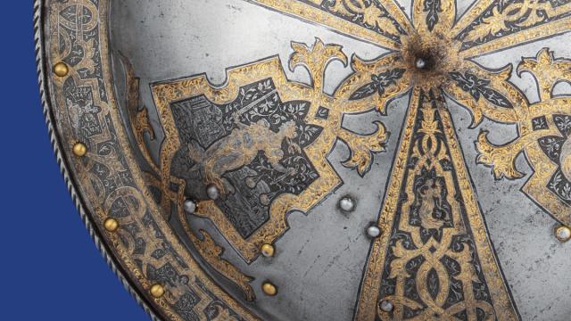 Ornate silver shield with intricate gold vine patterns and medieval animal motifs, accented by brass rivets on a blue background.