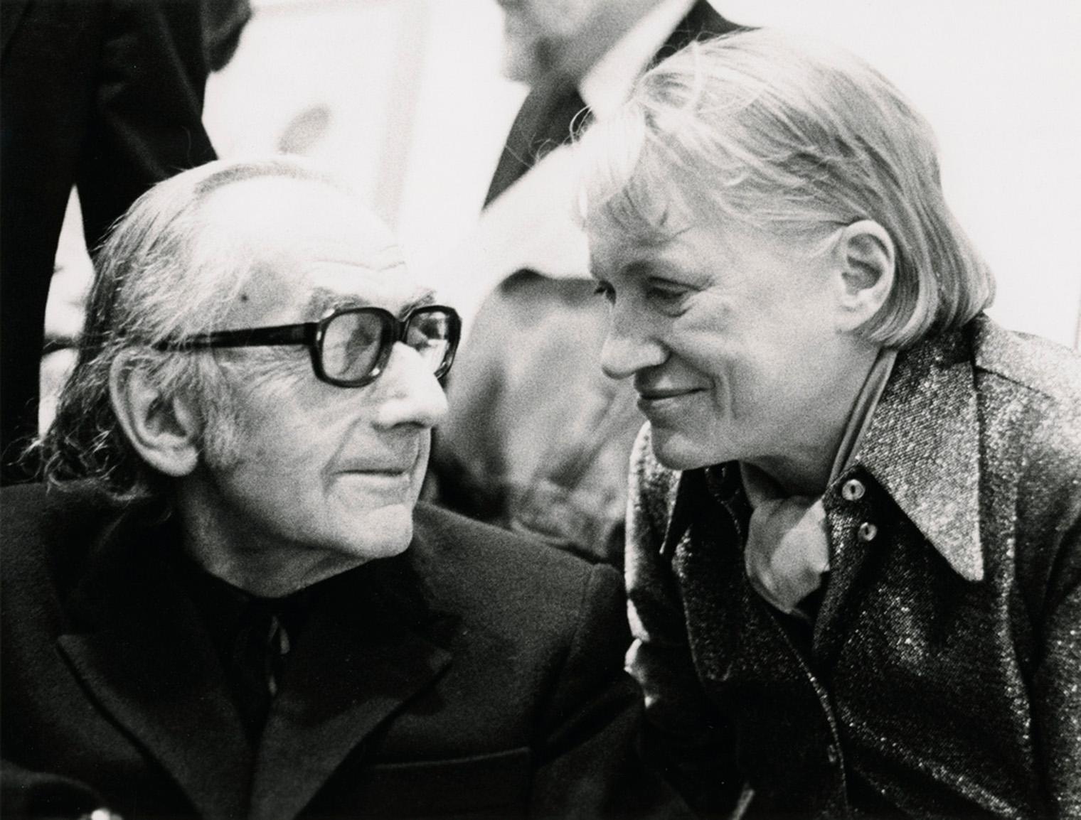 Two people share a warm moment. One, wearing glasses, looks towards the other with affection. The other smiles warmly, leaning closer. Black and white photo.