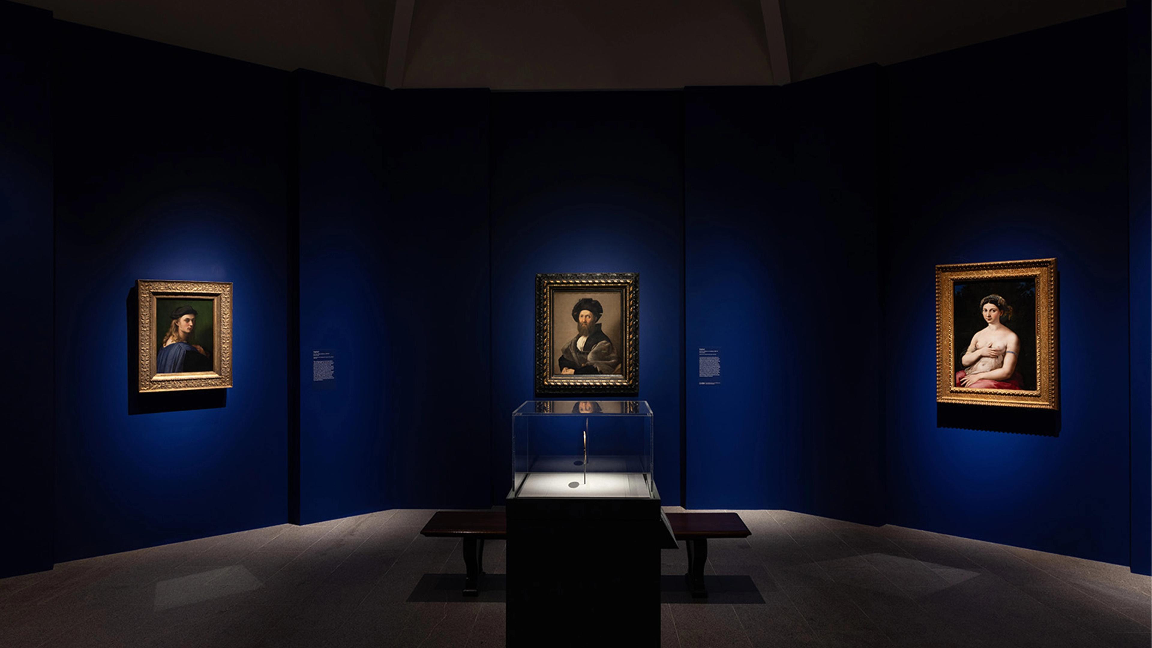 A dimly lit art gallery with deep blue walls displays three framed portraits.