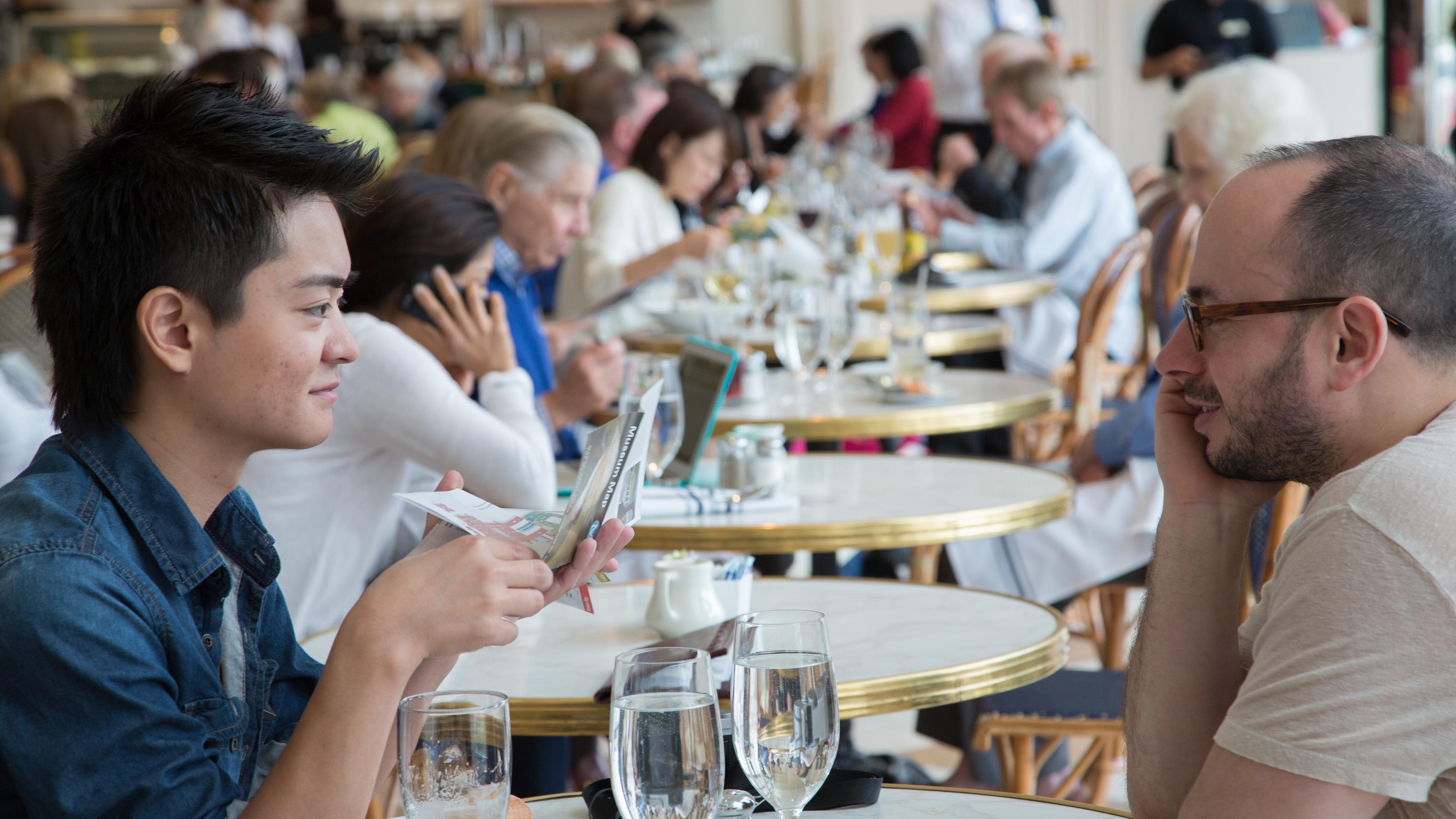 Two Musuem visitors sit at a table in the American Wing Cafe in The Met, talking over drinks.