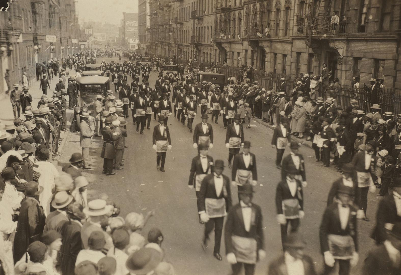 Vintage 1920s photo of a crowd of onlookers and a parade of suited men making their way down a street in Harlem