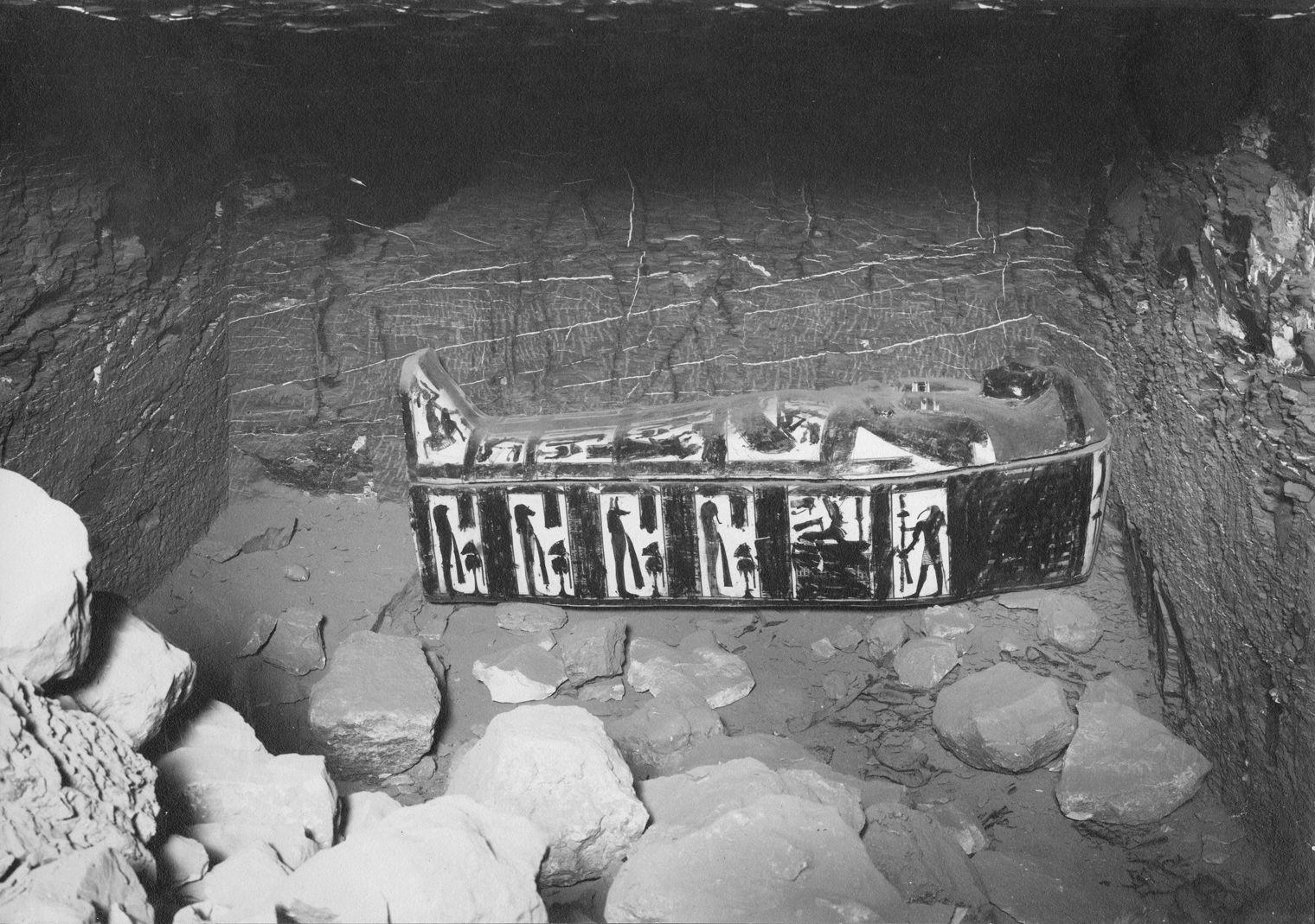 Black and white image of a rock cut chamber with a painted wooden coffin lying down in the back