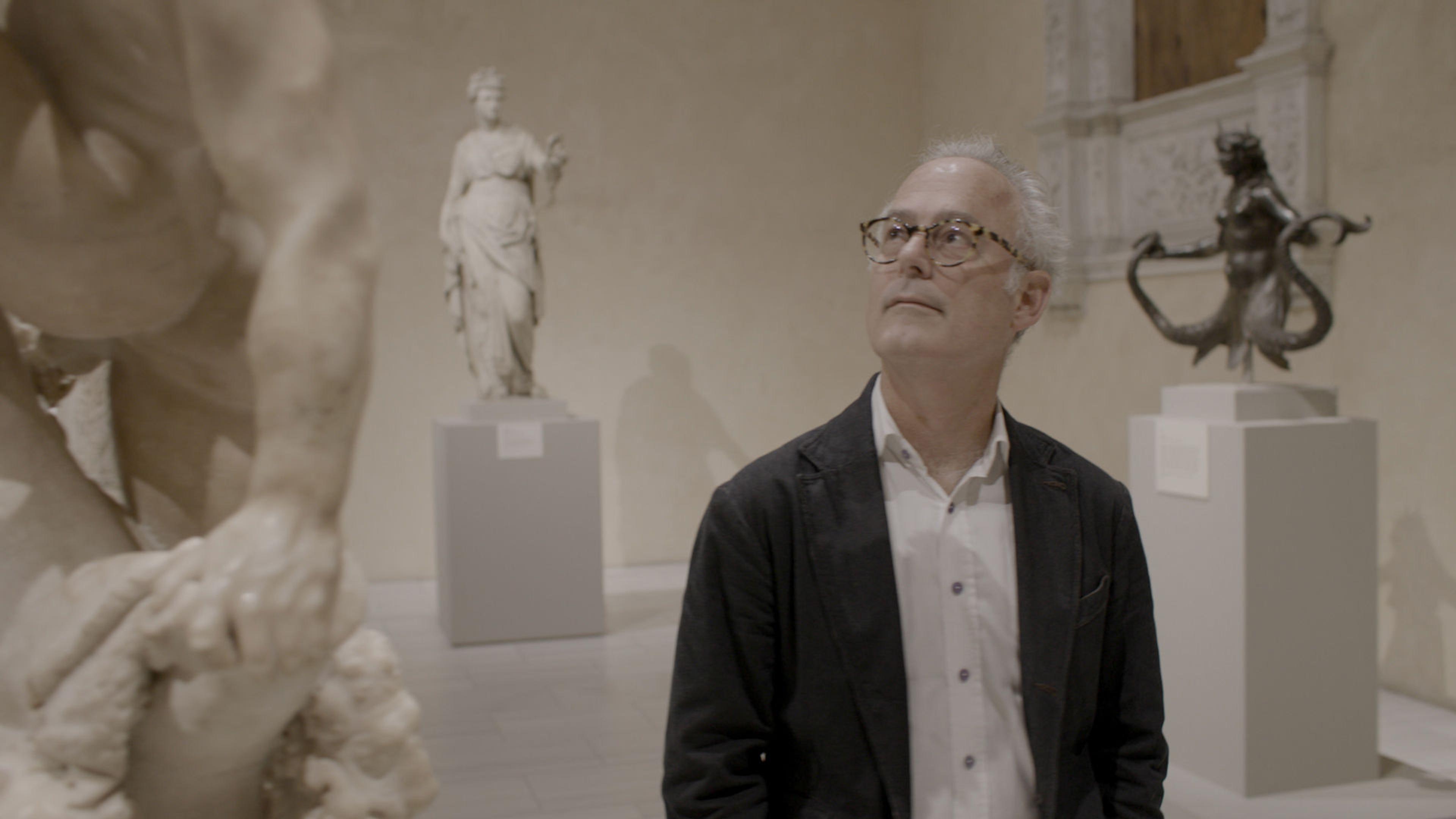 Writer Amor Towles looks upwards to his right at a statue. Two other statues, set on pedestals, are visible in the background