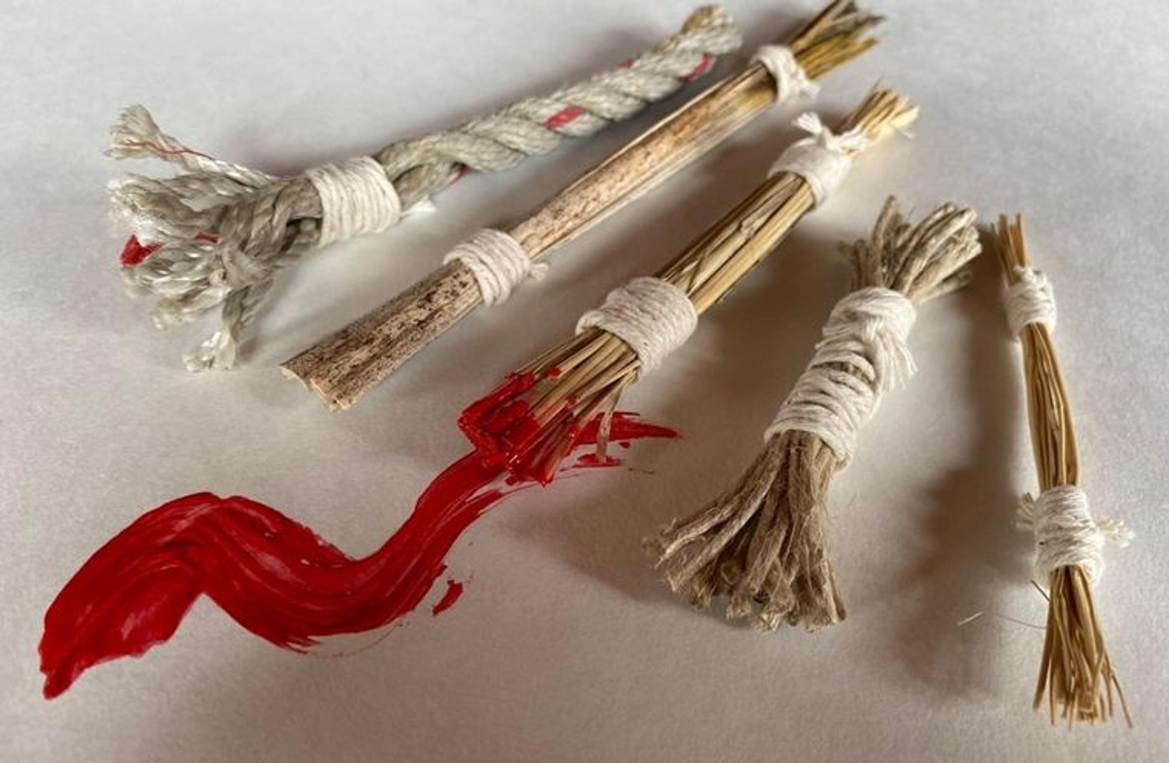 Five handmade paintbrushes built from materials like twigs and cord. One has red paint on the tip, leading to a zig-zag of red paint