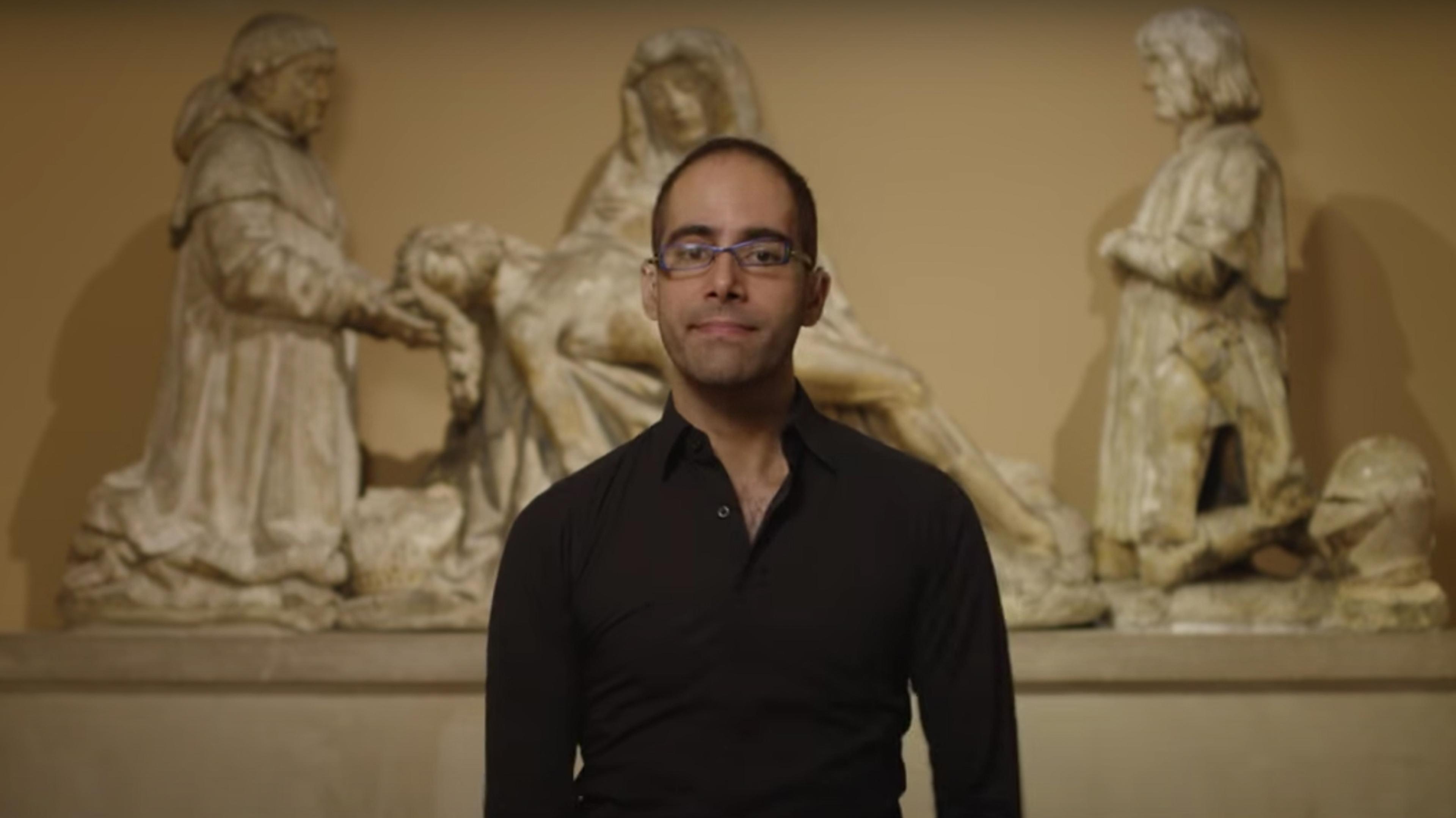 Art Historian and Met educator Emmanuel von Schack appears in front of a sculpture depicting figures encircling and mourning a deceased person laying in shrouds in the center of the figural group