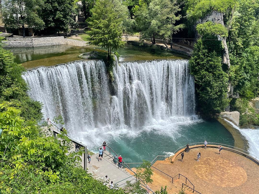 The Pliva Waterfall pouring into the Vrbas below the green cliffs of Jajce, with the viewing platform on the right