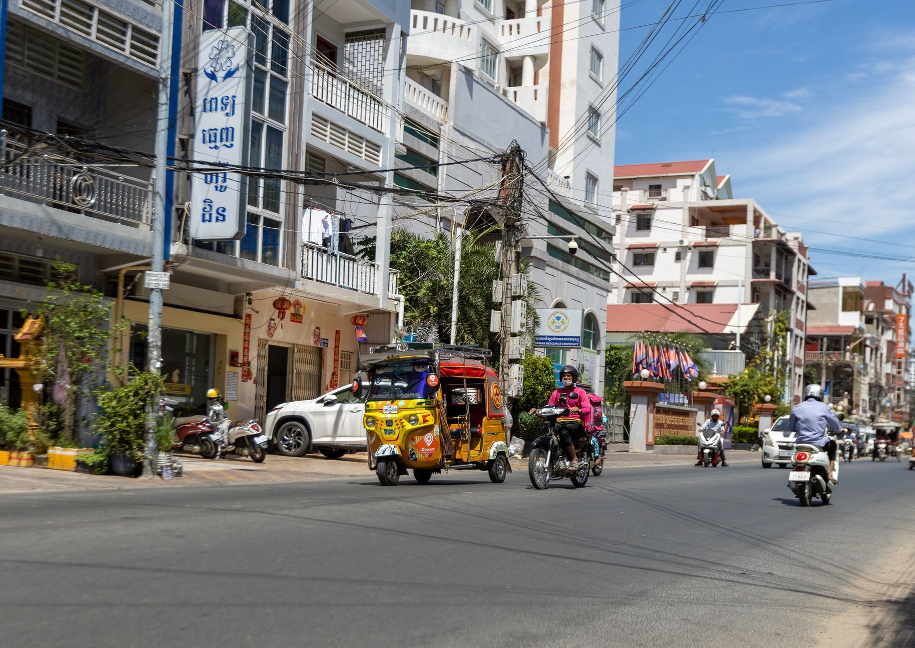 A tuk-tuk and motorbikes moving through a busy street in Phnom Penh past small shopfronts