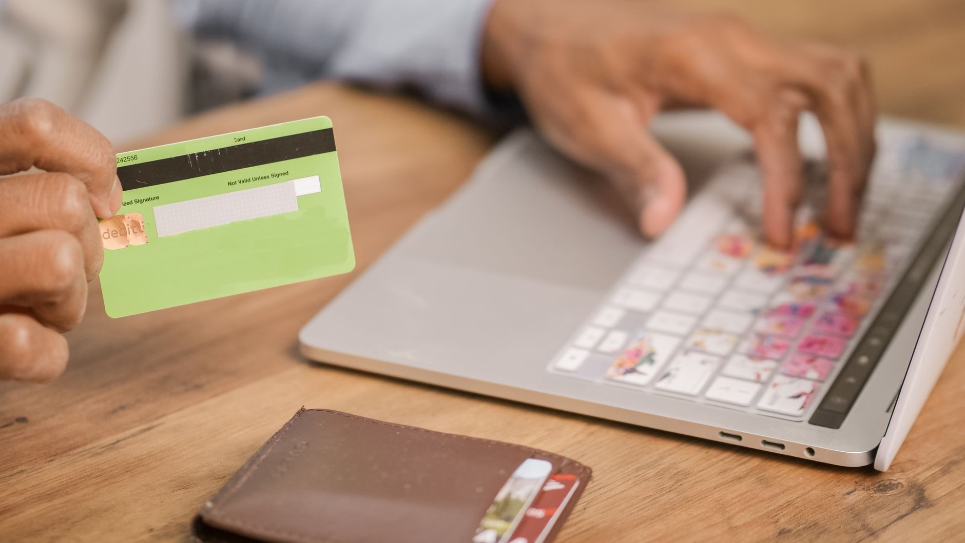 Hand holding a payment card beside an open laptop and a wallet on a desk