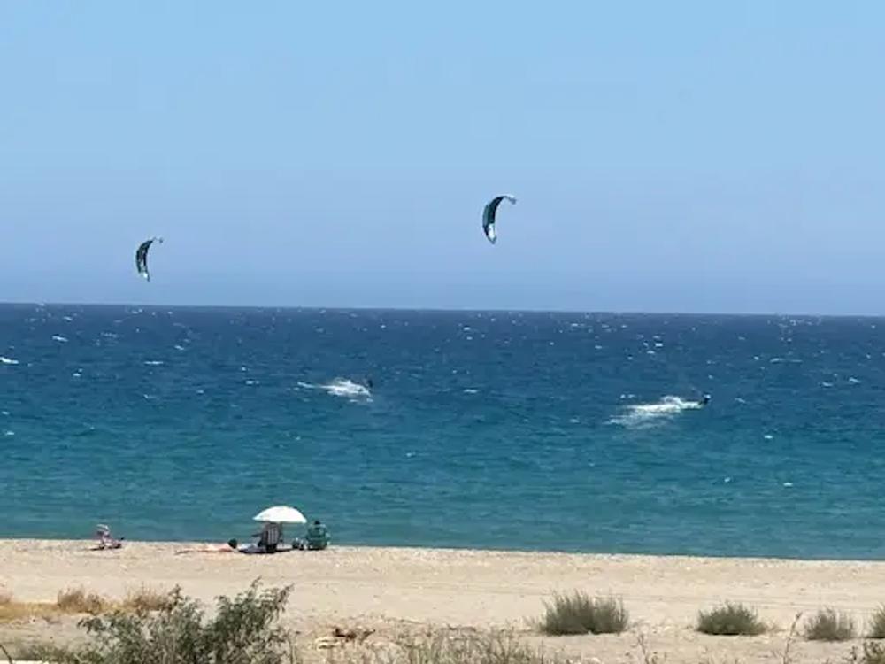 Clear Blue sky - interrupted by 2 Kite surfers, a darker blue sea - with the splashes made by the surf and the yellow of the sand on the beach with people sitting under a white umbrella.