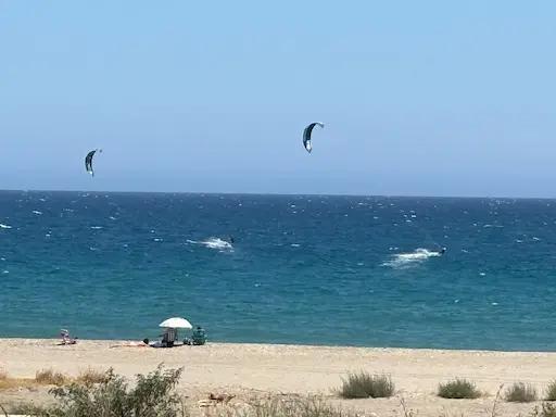 Clear Blue sky - interrupted by 2 Kite surfers, a darker blue sea - with the splashes made by the surf and the yellow of the sand on the beach with people sitting under a white umbrella.