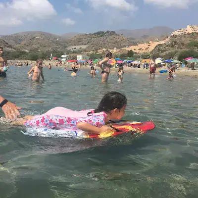 A child friendly beach in Mojácar in southern Spain, Showing a girl in a pink sun protection swimming costume, laying on a body board, with people in the background standing in the water. Hills in the background.