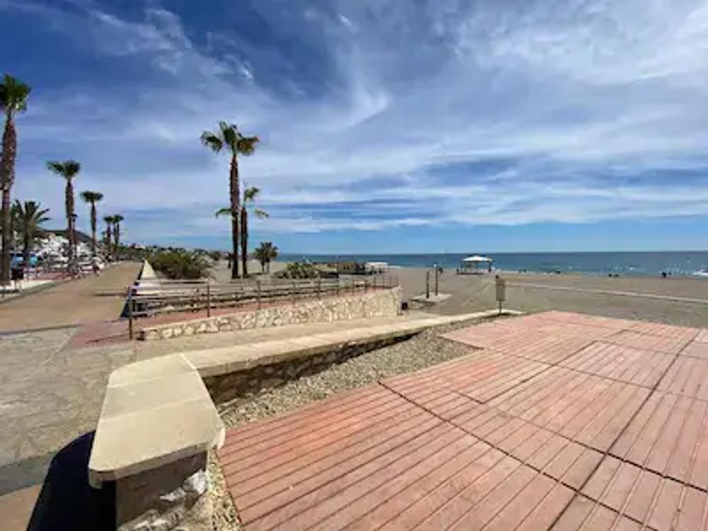 Accessible beach in Mojacar.  The paseo which is above sea level is shown on the left, has a concrete slope down to beach level that turns into a wooden "boardwalk" across the sand towards the shore.  There is also a fixed shelter providing shade.