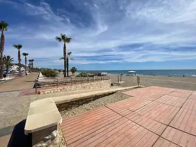 Accessible beach in Mojacar. The paseo which is above sea level is shown on the left, has a concrete slope down to beach level that turns into a wooden "boardwalk" across the sand towards the shore. There is also a fixed shelter providing shade.