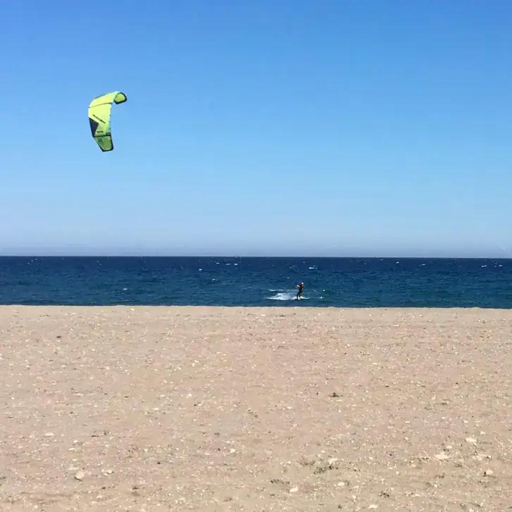 A beach in Mojácar with a kitesurfer with a green kite. The sky is blue, the sea is a deeper blue and the sand in the foreground has some small pebbles, but is mainly sand.