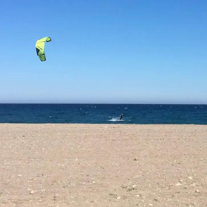 A beach in Mojácar with a kitesurfer with a green kite. The sky is blue, the sea is a deeper blue and the sand in the foreground has some small pebbles, but is mainly sand.