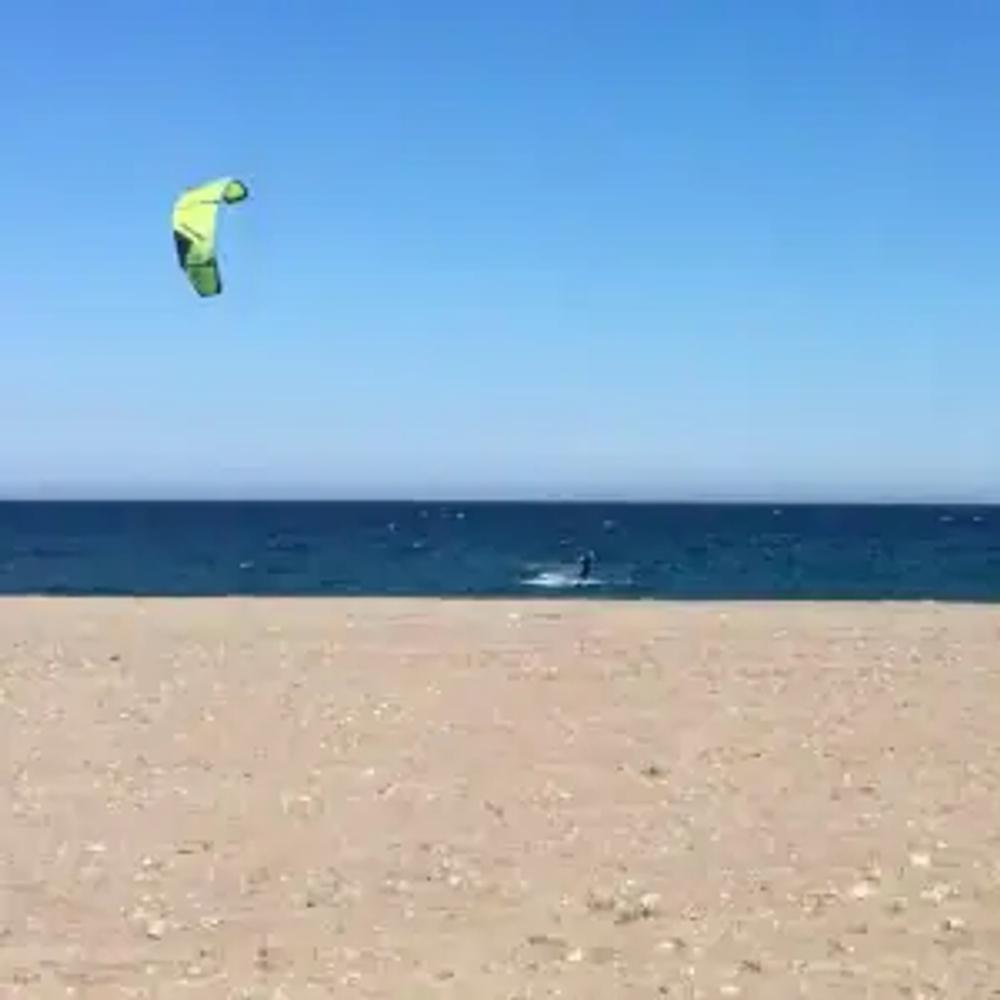 A beach in Mojácar with a kitesurfer with a green kite. The sky is blue, the sea is a deeper blue and the sand in the foreground has some small pebbles, but is mainly sand.