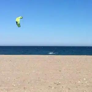 A beach in Mojácar with a kitesurfer with a green kite. The sky is blue, the sea is a deeper blue and the sand in the foreground has some small pebbles, but is mainly sand.