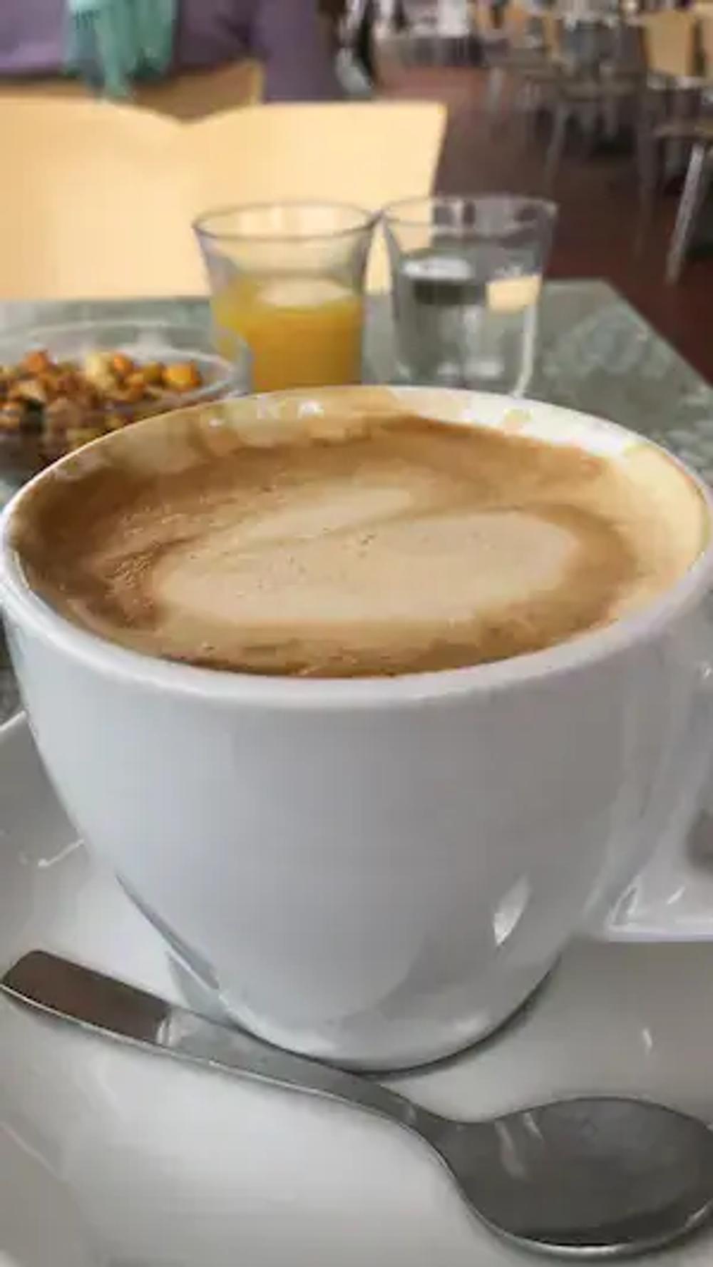 Portrait photo of a cafe con leche and faded in the background a small bowl of nuts, and two small glasses. One with juice and one with water. Taking expensive real estate if you're trying to work.