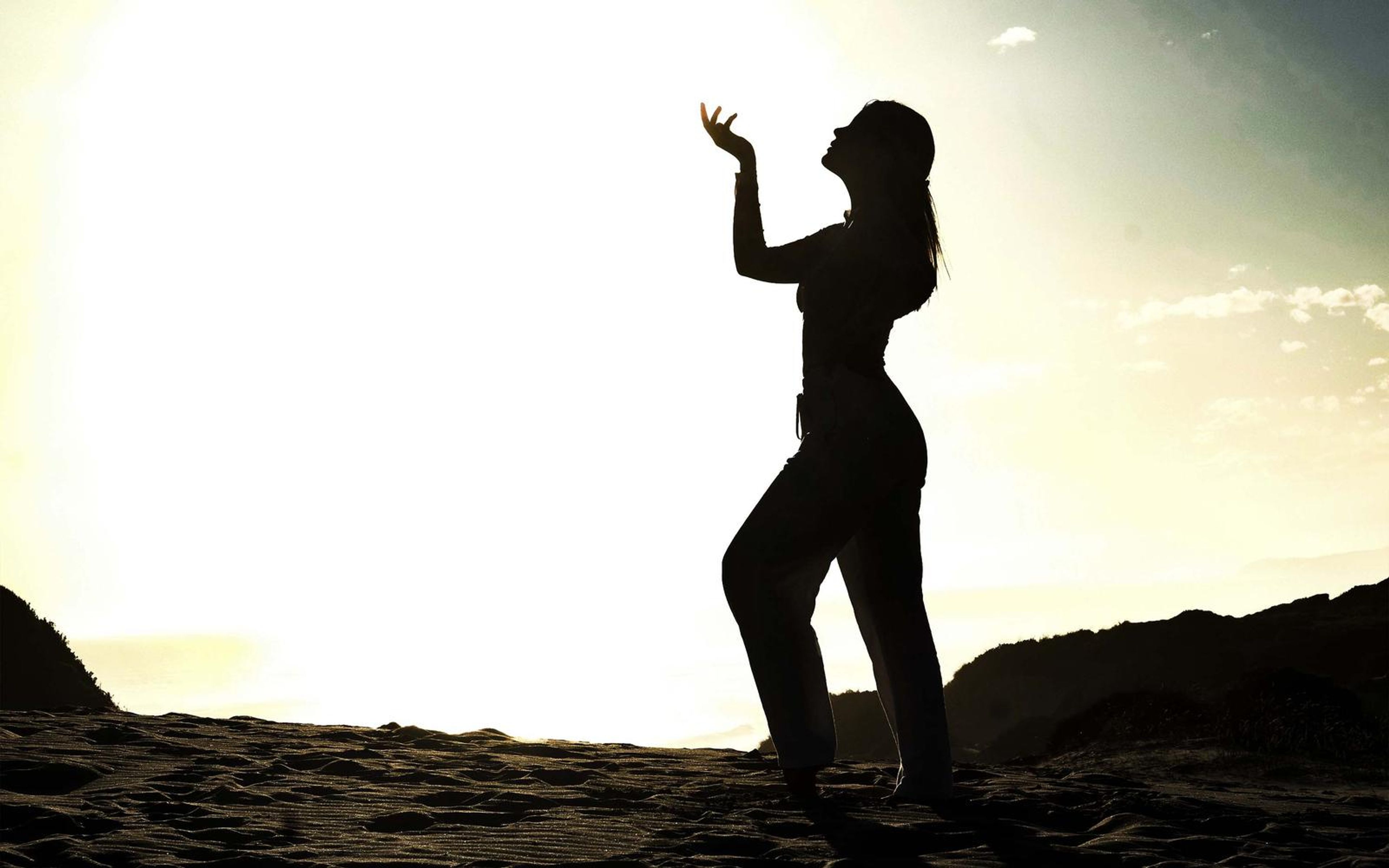 Silhouette of a woman with one hand raised against a bright sky above dunes