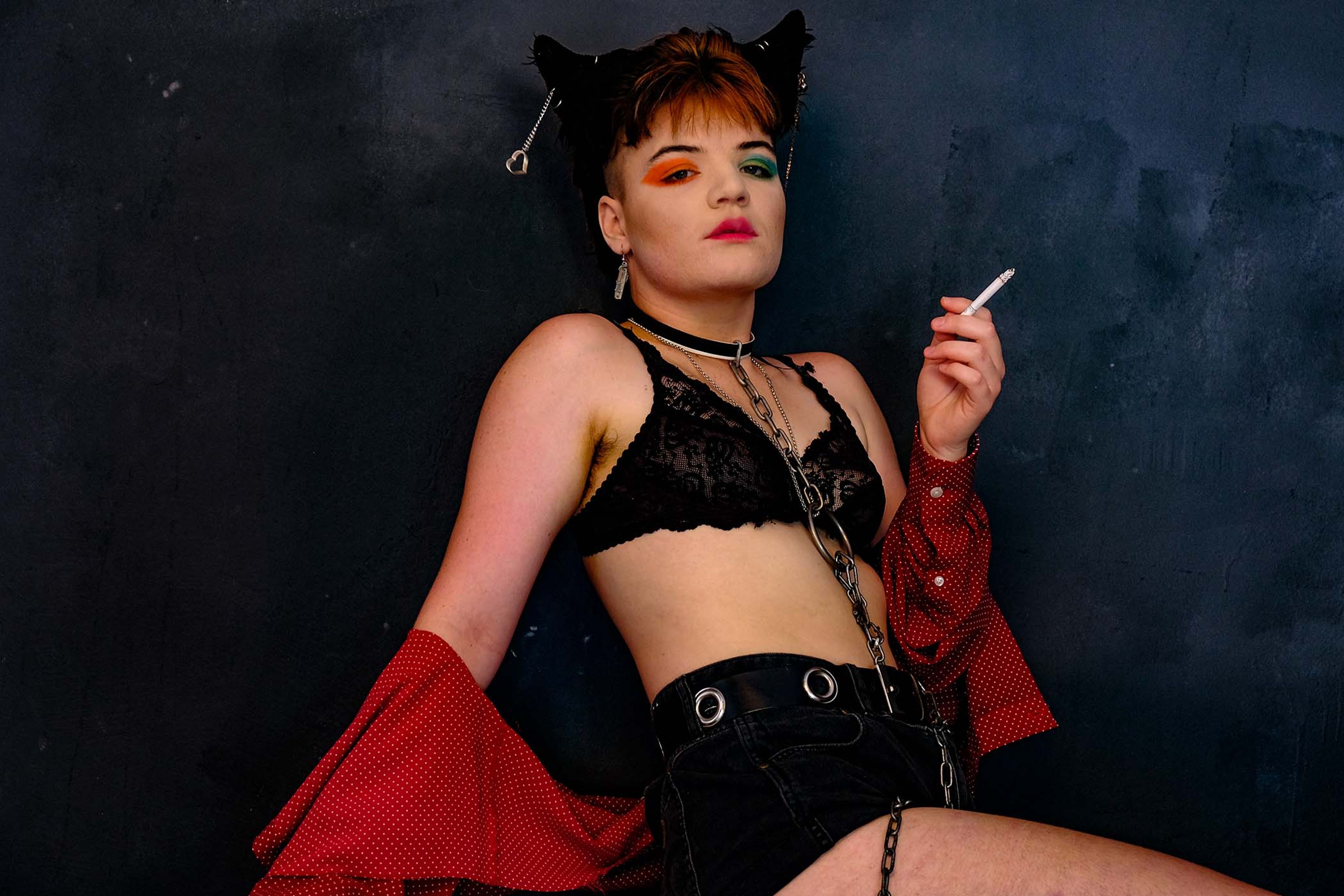 Portrait, subject in a lace bra and cat-ear headpiece holding a cigarette, red cardigan draped off the shoulders